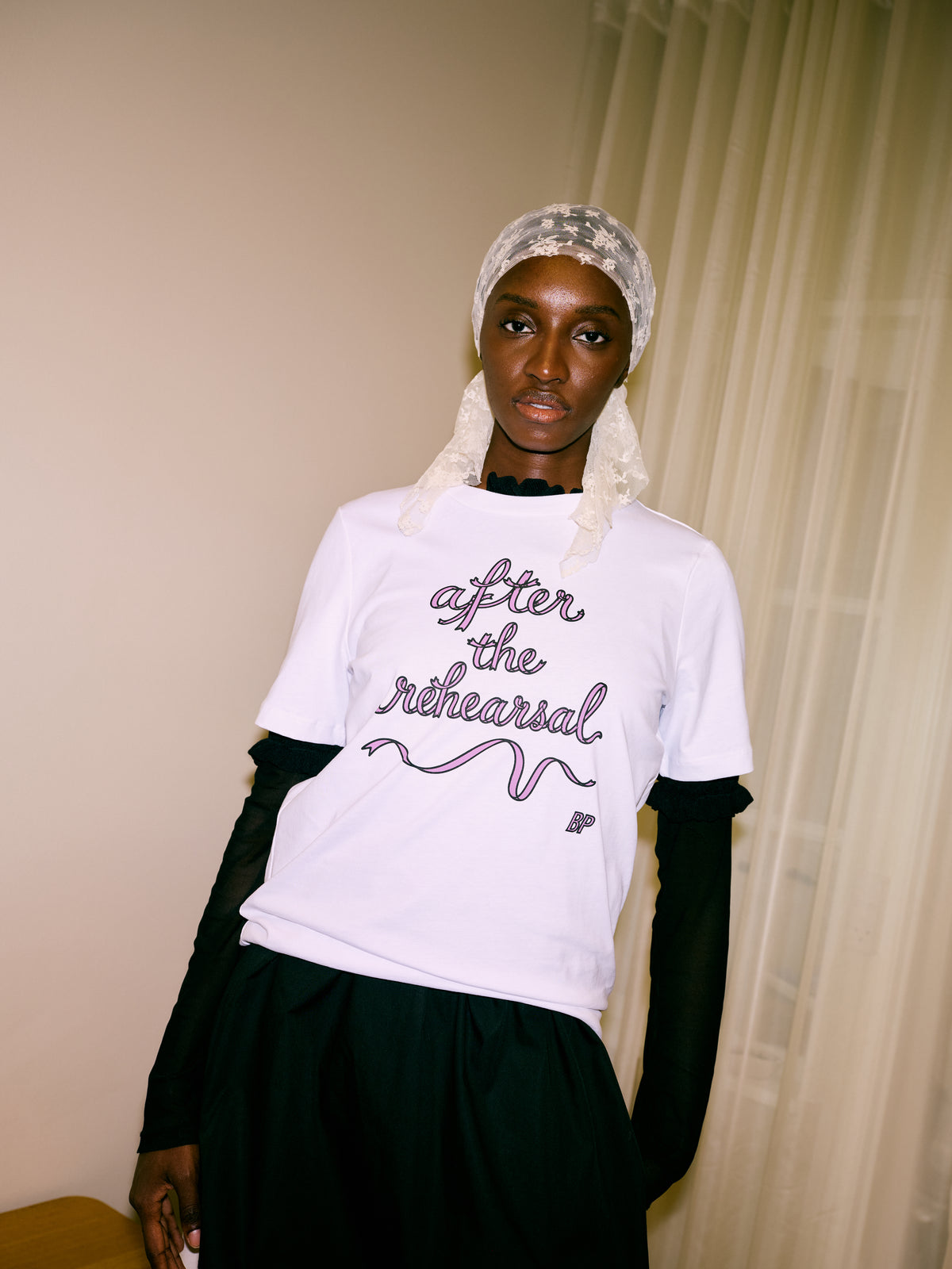 Gecropt campagneportret van een model in een wit T-shirt met roze en zwarte letters waarop 'Na de repetitie,' staat, en een crèmekleurige kanten hoofddoek.