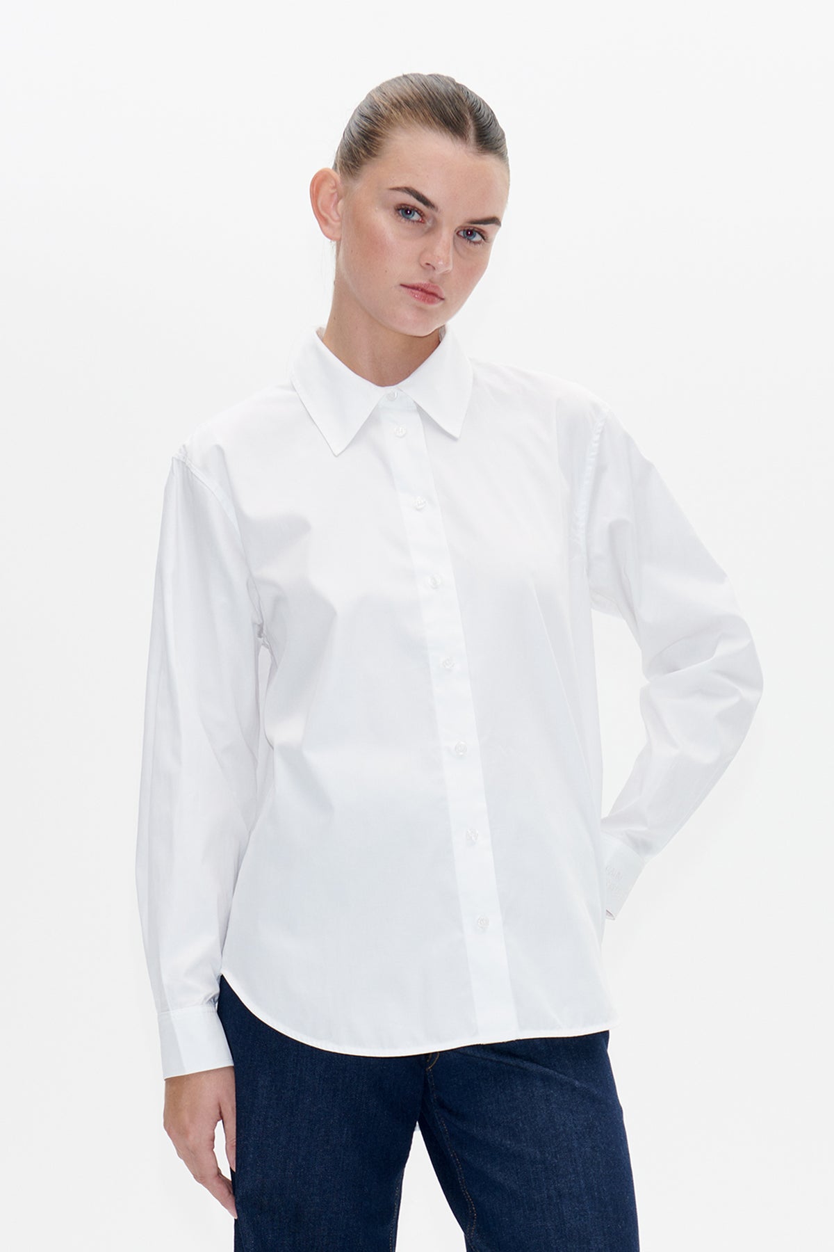 Model wearing a white long-sleeved button-front shirt with a pointed collar, paired with dark indigo jeans.