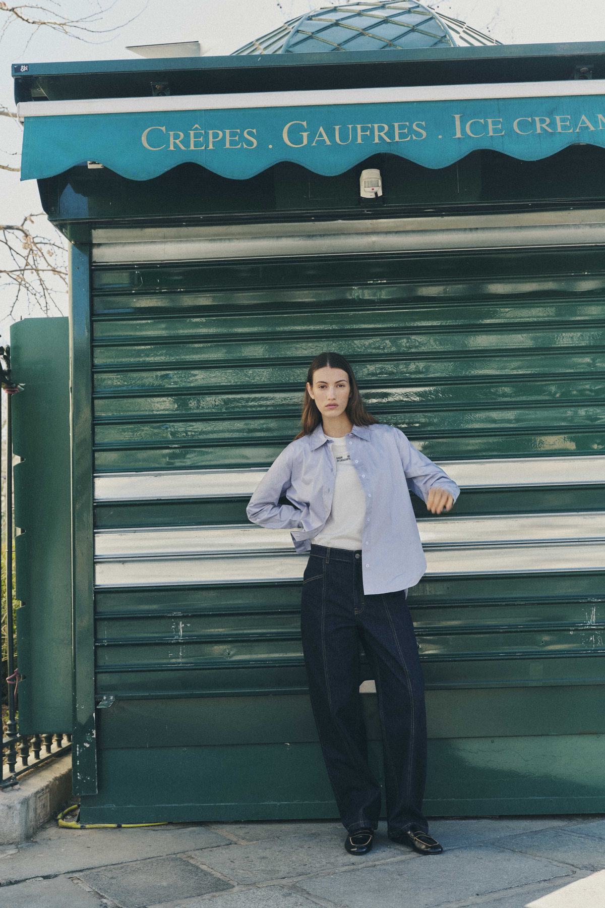 Model in a pale blue button-front shirt over a white T-shirt and dark wide-leg jeans, standing before a green storefront