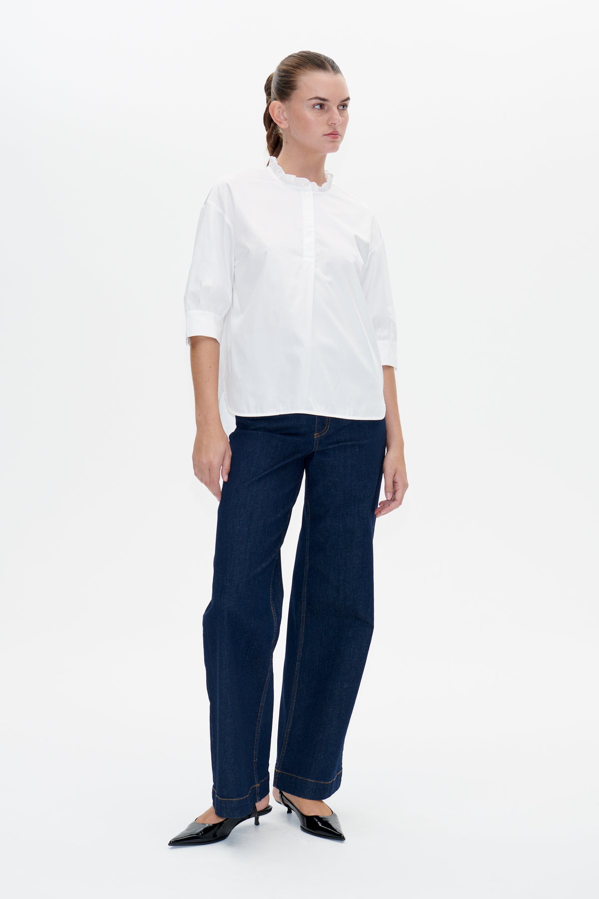 Model trägt eine weiße Bluse mit Rüschenkragen und Dreiviertelärmeln, kombiniert mit dunkelblauen Jeans.
