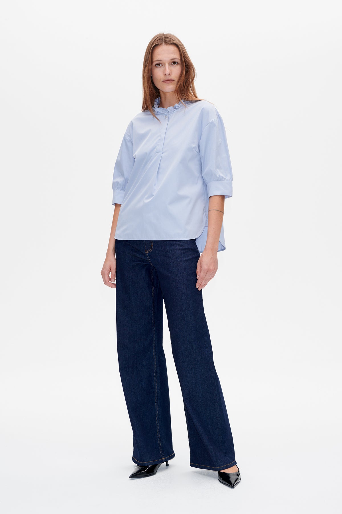 Front view of a pale blue blouse with a ruffled collar, button placket and elbow-length sleeves, paired with dark wide-leg jeans.