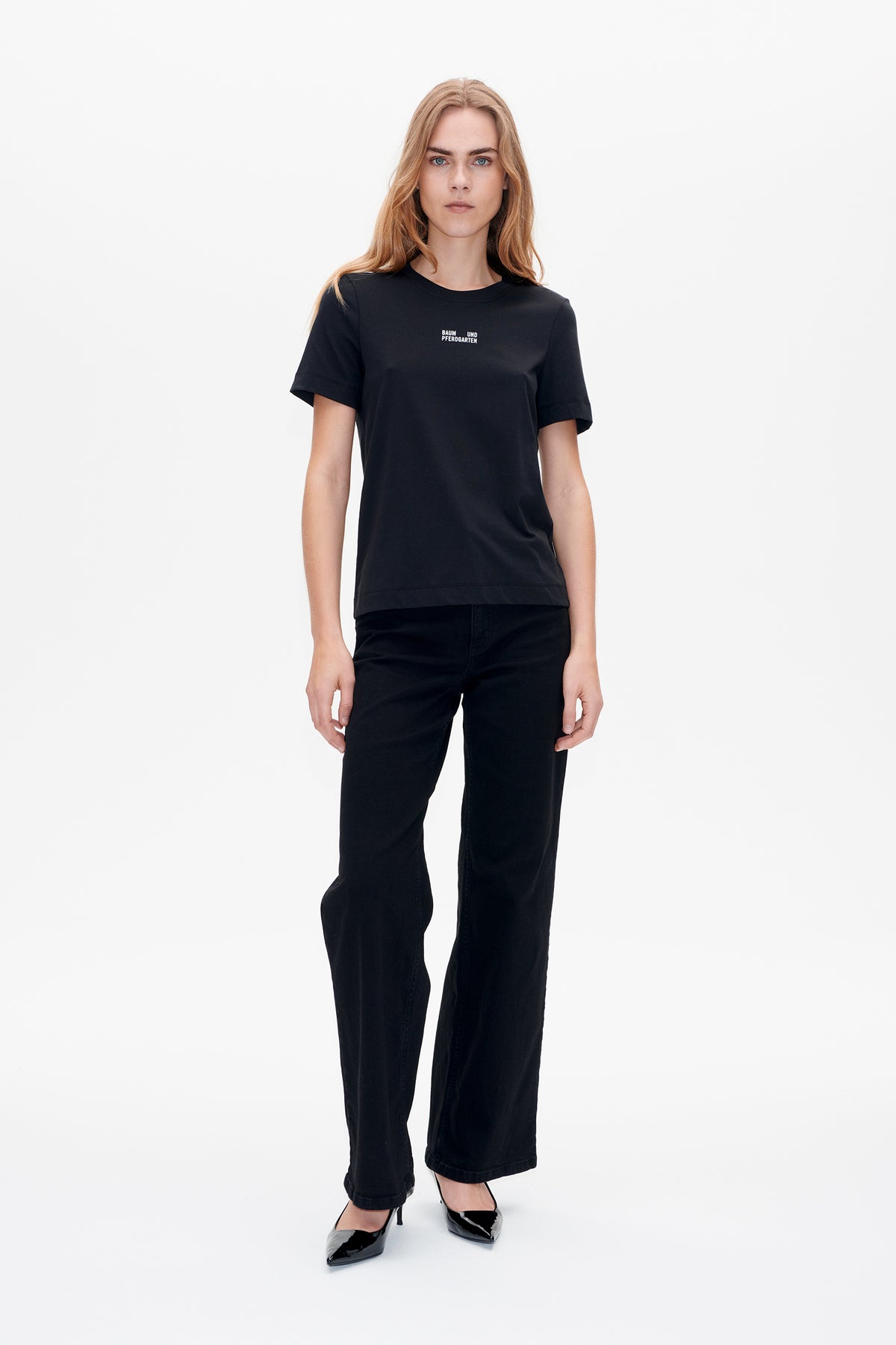 Vue de face d'un mannequin portant un T-shirt noir à manches courtes et col rond, orné d'un petit logo blanc Baum und Pferdgarten sur la poitrine et un jean noir à jambes larges.