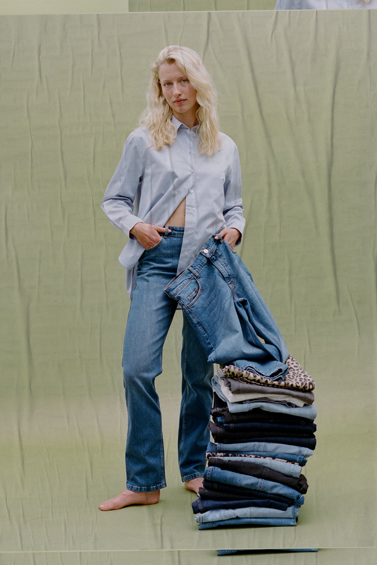 Model in een lichtblauwe blouse met knopen en blauwe jeans houdt een denimstuk naast opgevouwen kleding tegen een zachtgroene achtergrond.