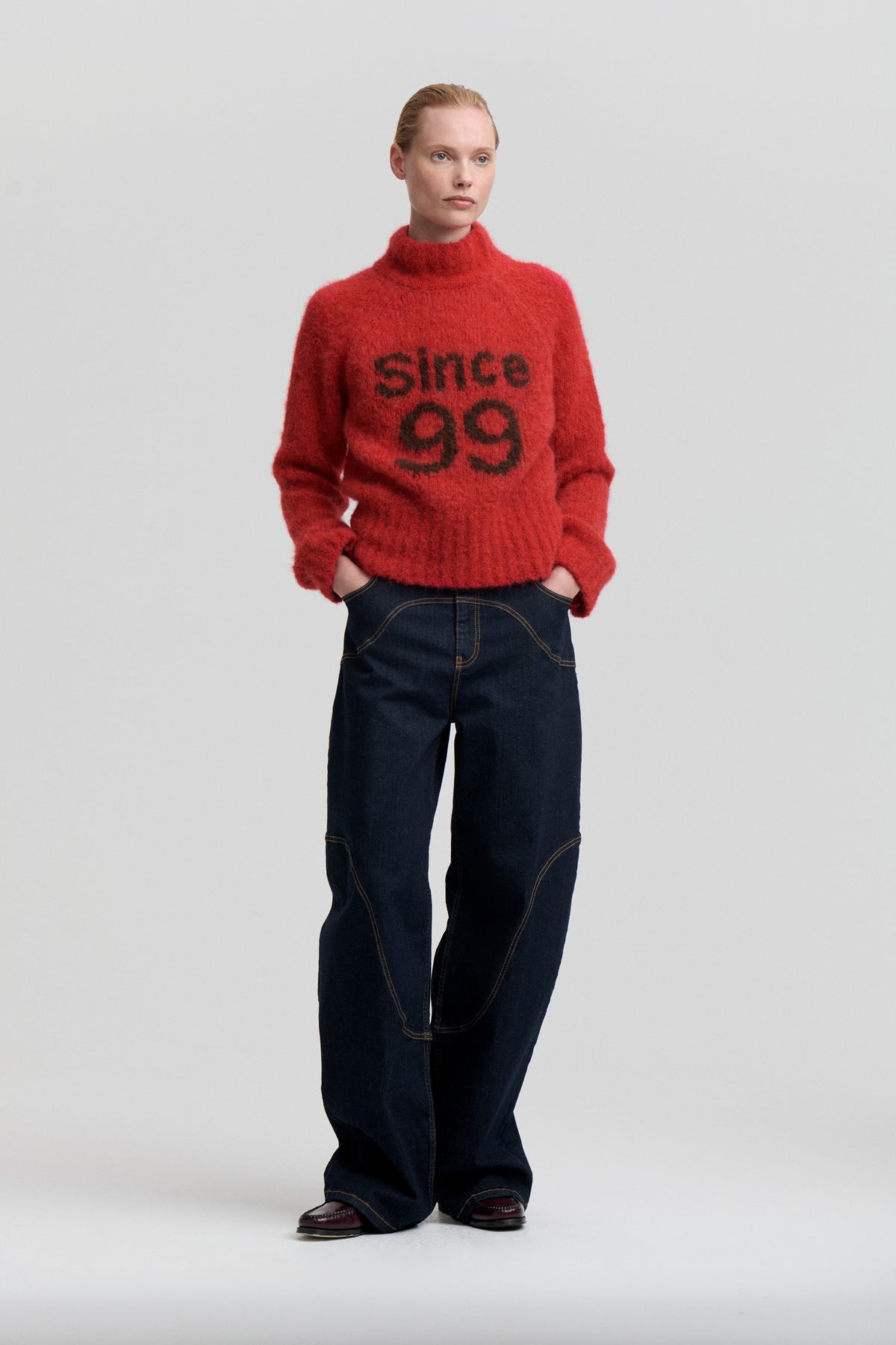Model wearing a fuzzy red high-neck sweater with black “since 99” lettering and dark indigo wide-leg jeans against a pale studio backdrop.