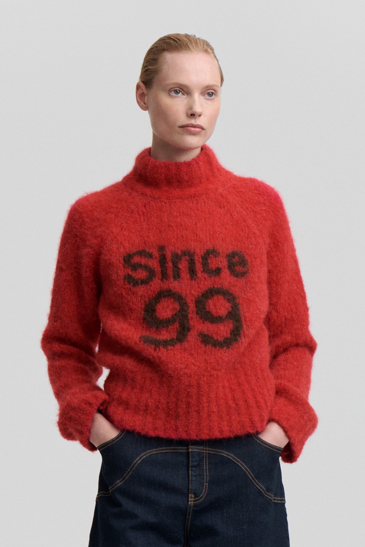 Close-up of a model wearing a fuzzy red high-neck sweater with bold black lettering and a graphic face motif.