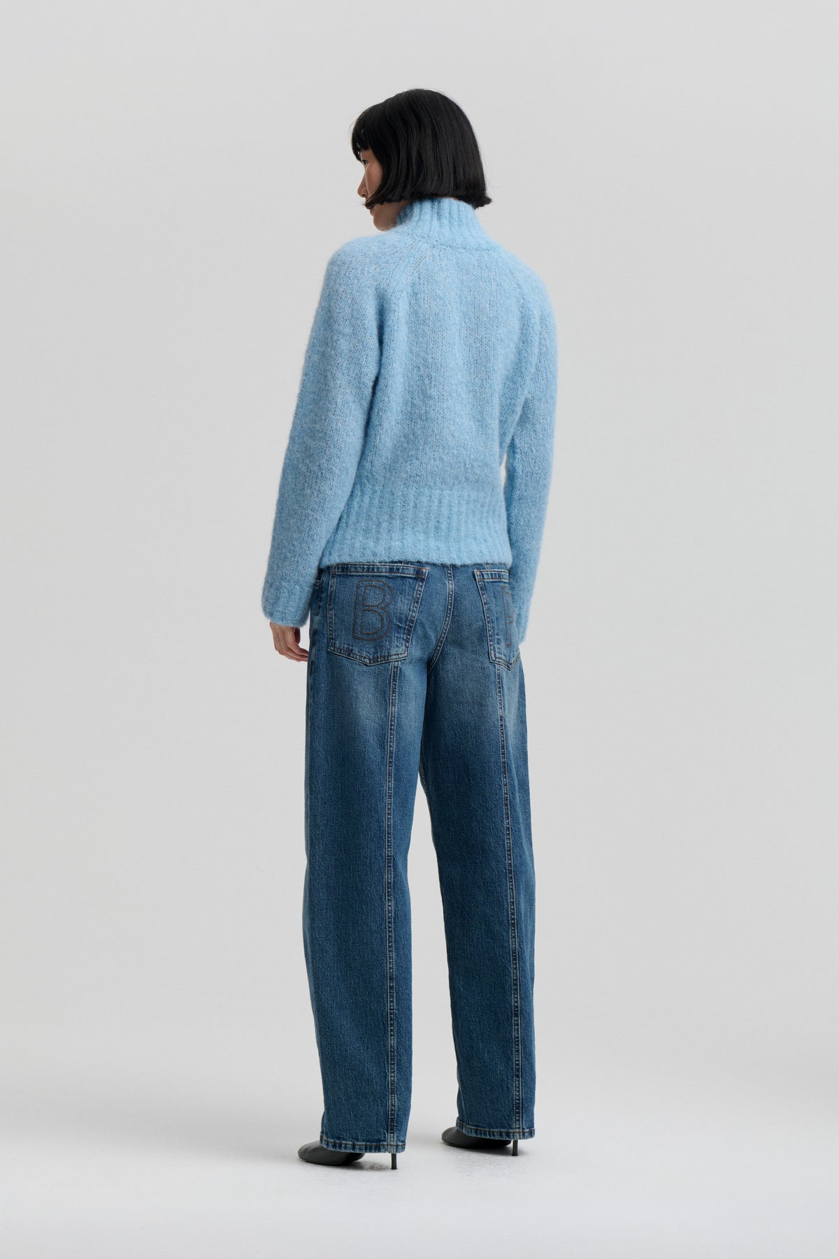 Back view of a model wearing a fuzzy light blue high-neck sweater and wide-leg blue jeans with outlined lettering on the back pockets.