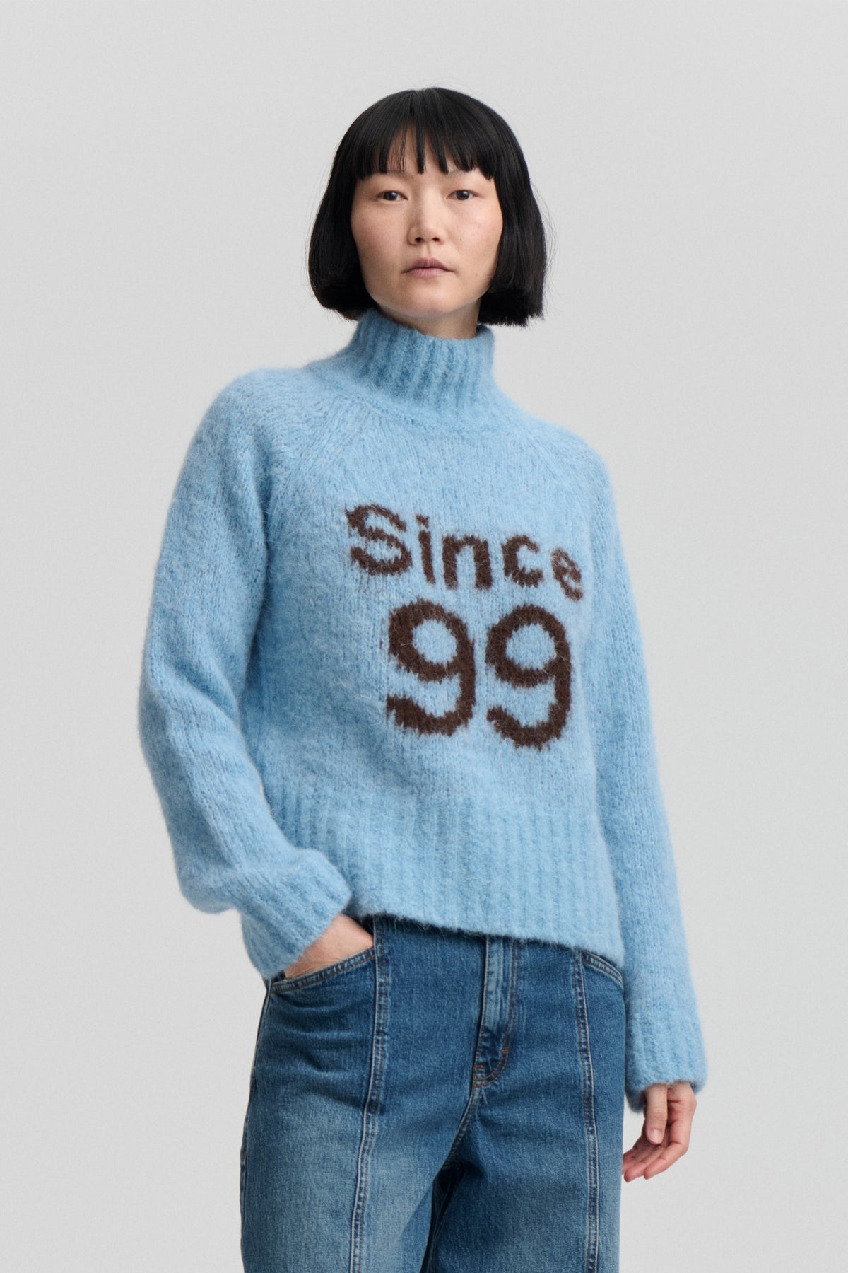 Model wearing a fuzzy light blue high-neck sweater with brown “STAY COOL” lettering and blue jeans, shown from the front.