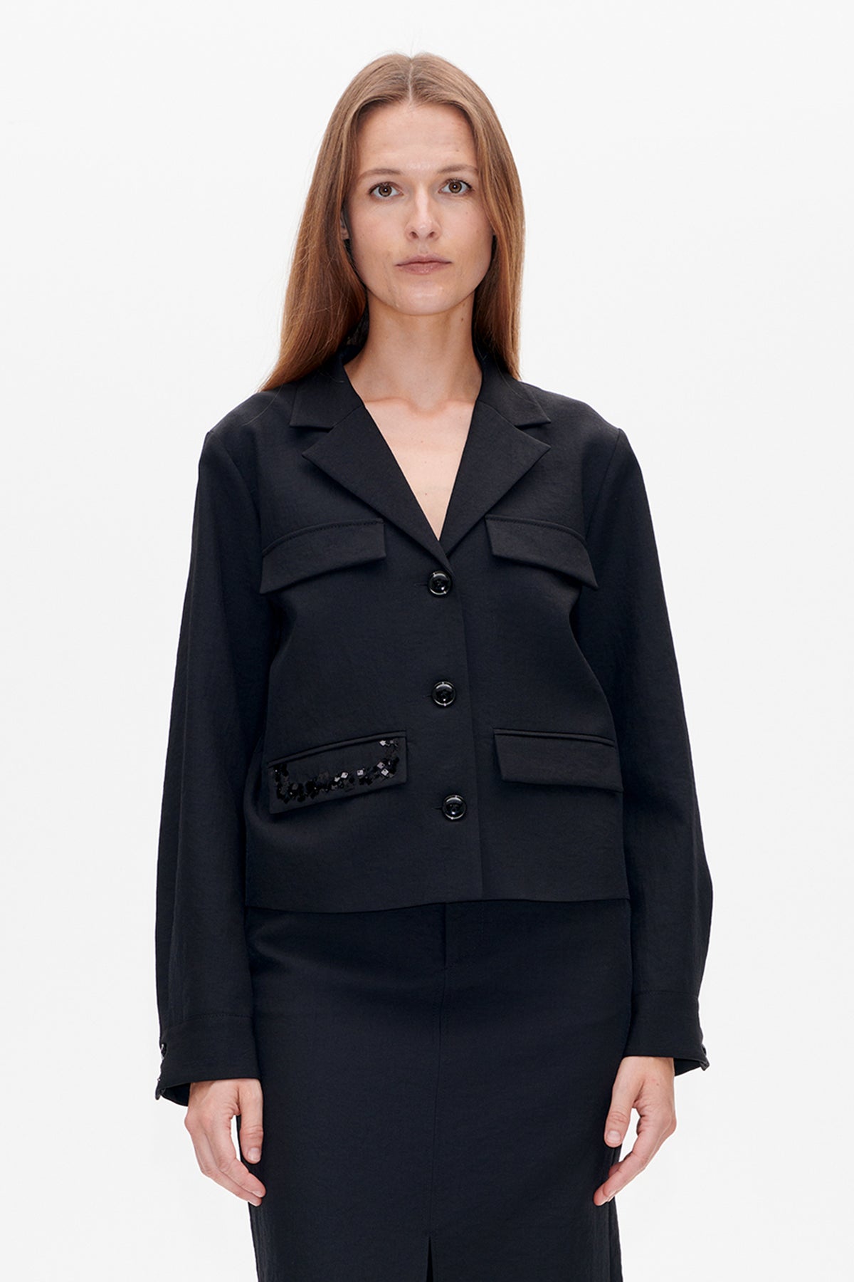 Vooraanzicht van een zwarte lange blazer met ingesneden revers, knoopsluiting, klepzakken en pailletten op één klepzak.