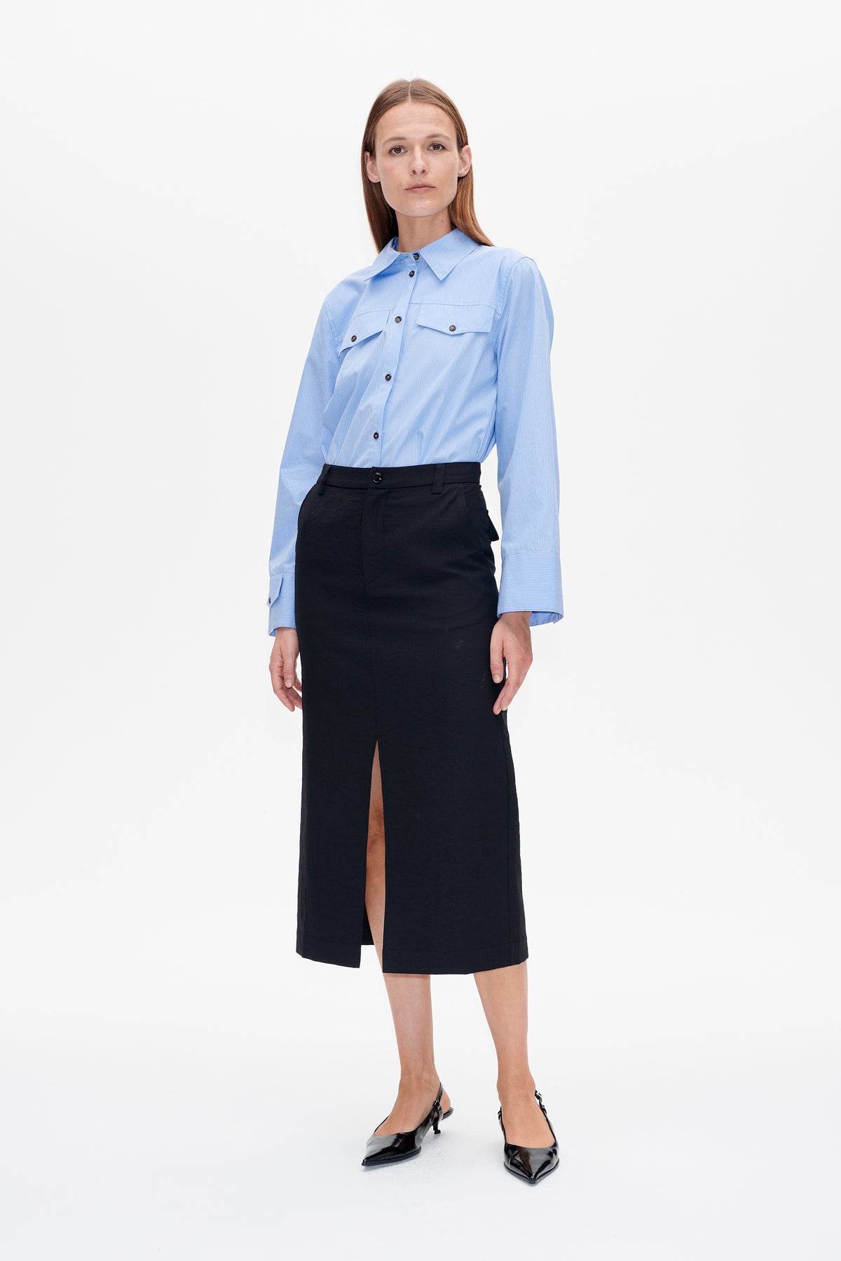 Front view of a model wearing a light blue pinstriped button-up shirt with chest pockets and a black midi skirt with a front slit.