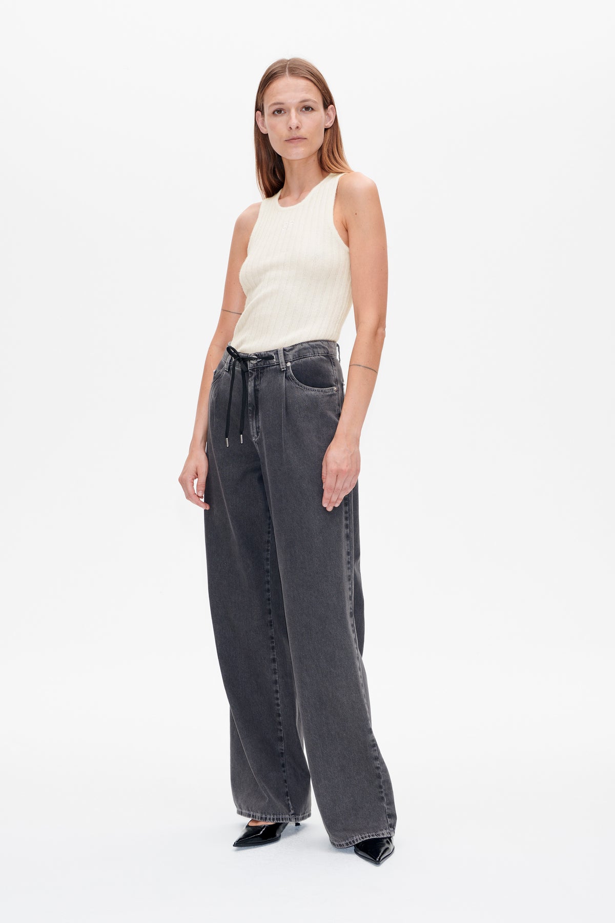 Vorderansicht eines Models, das ein elfenbeinfarbenes, geripptes ärmelloses Top, anthrazitfarbene Jeans mit weitem Bein und einen schwarzen Gürtel mit Kordelzug trägt.