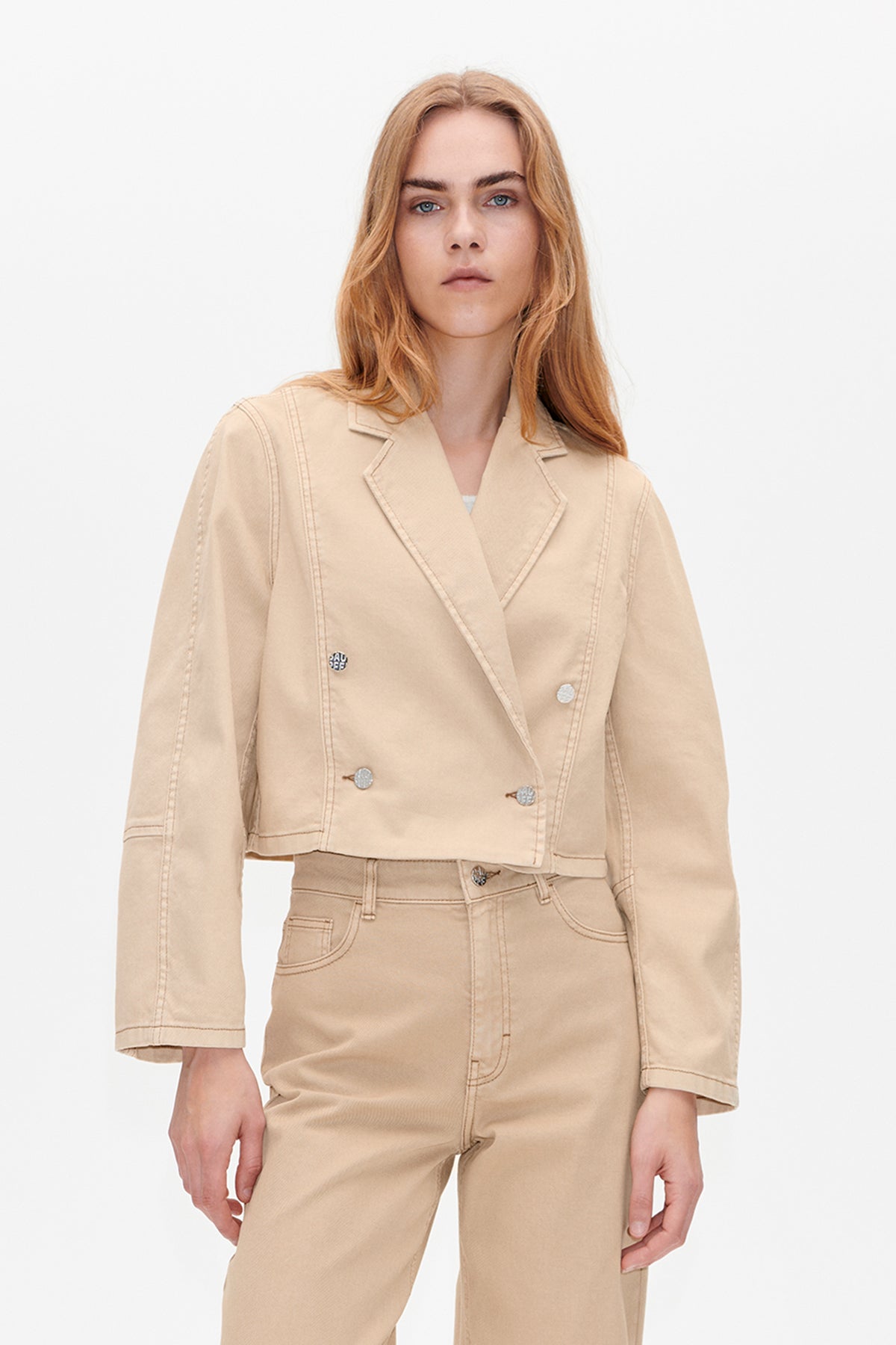 Front view of a model wearing a sand-beige cropped jacket with wide lapels, contrast topstitching and silver buttons, paired with matching trousers.