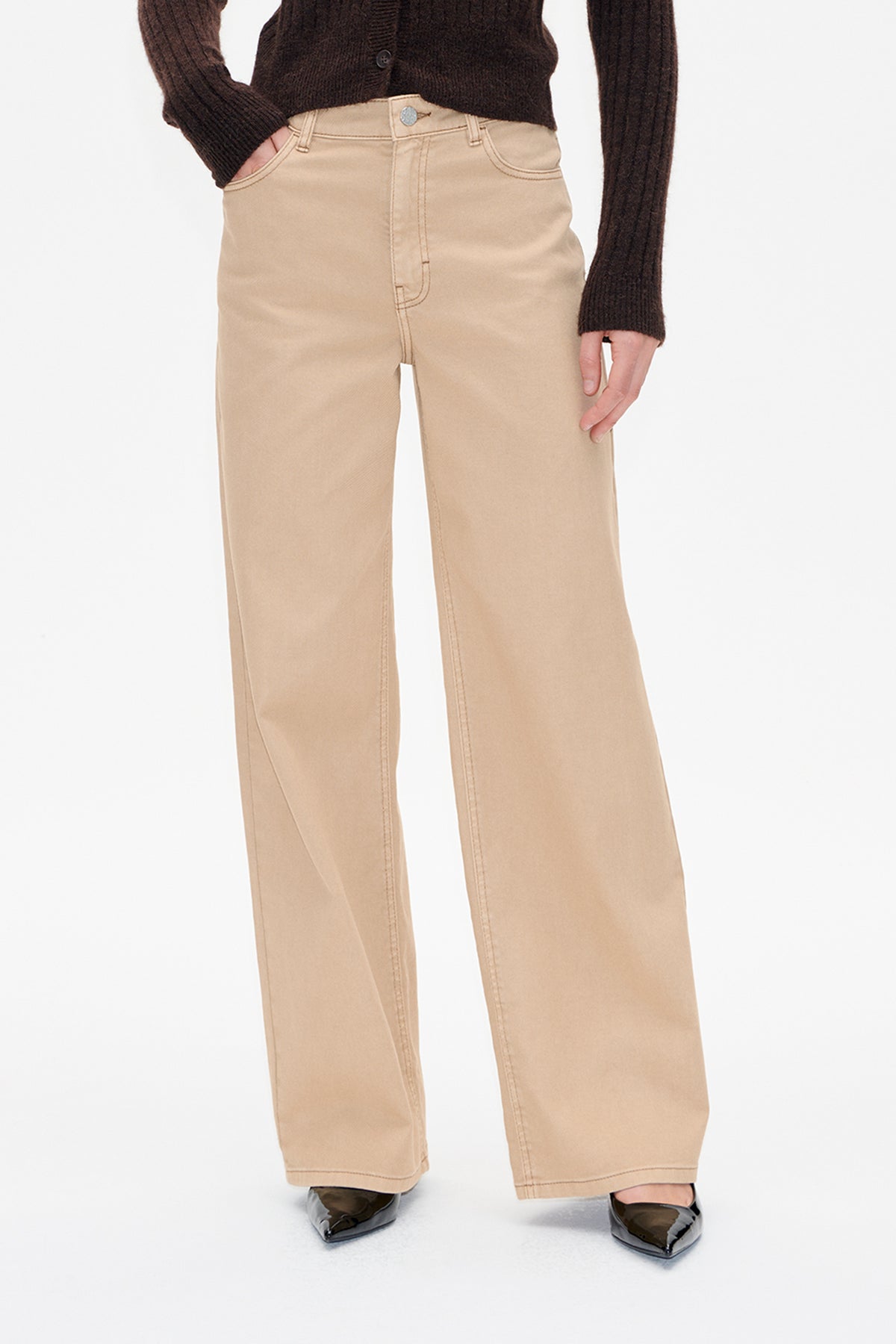 Close front view of beige wide-leg trousers with belt loops, button fastening and curved pockets, worn with a dark brown ribbed knit top.