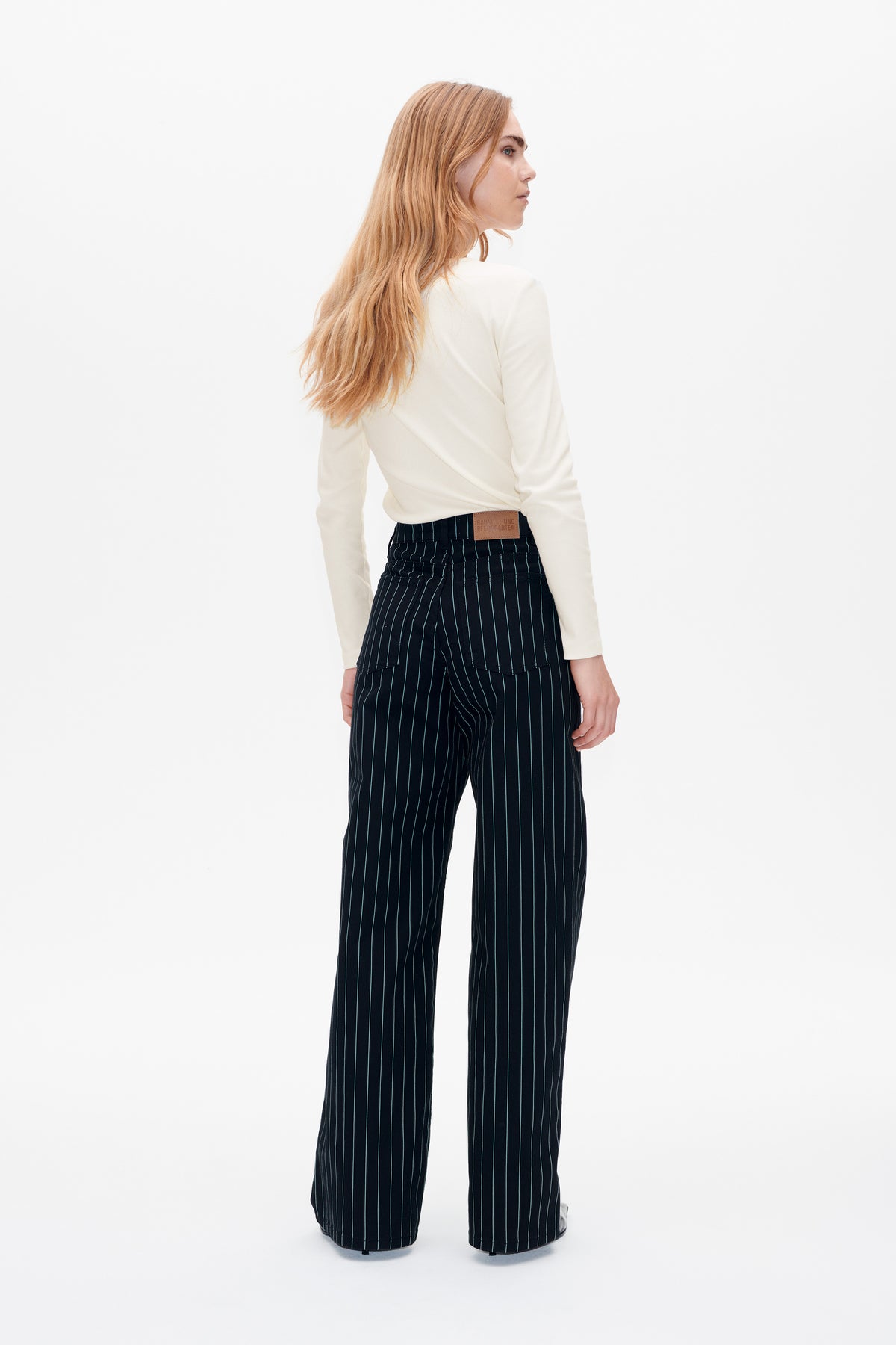 Model von hinten in einem cremefarbenen Langarmtop und einer schwarzen Nadelstreifen-Hose mit weitem Bein und aufgesetzten Gesäßtaschen.