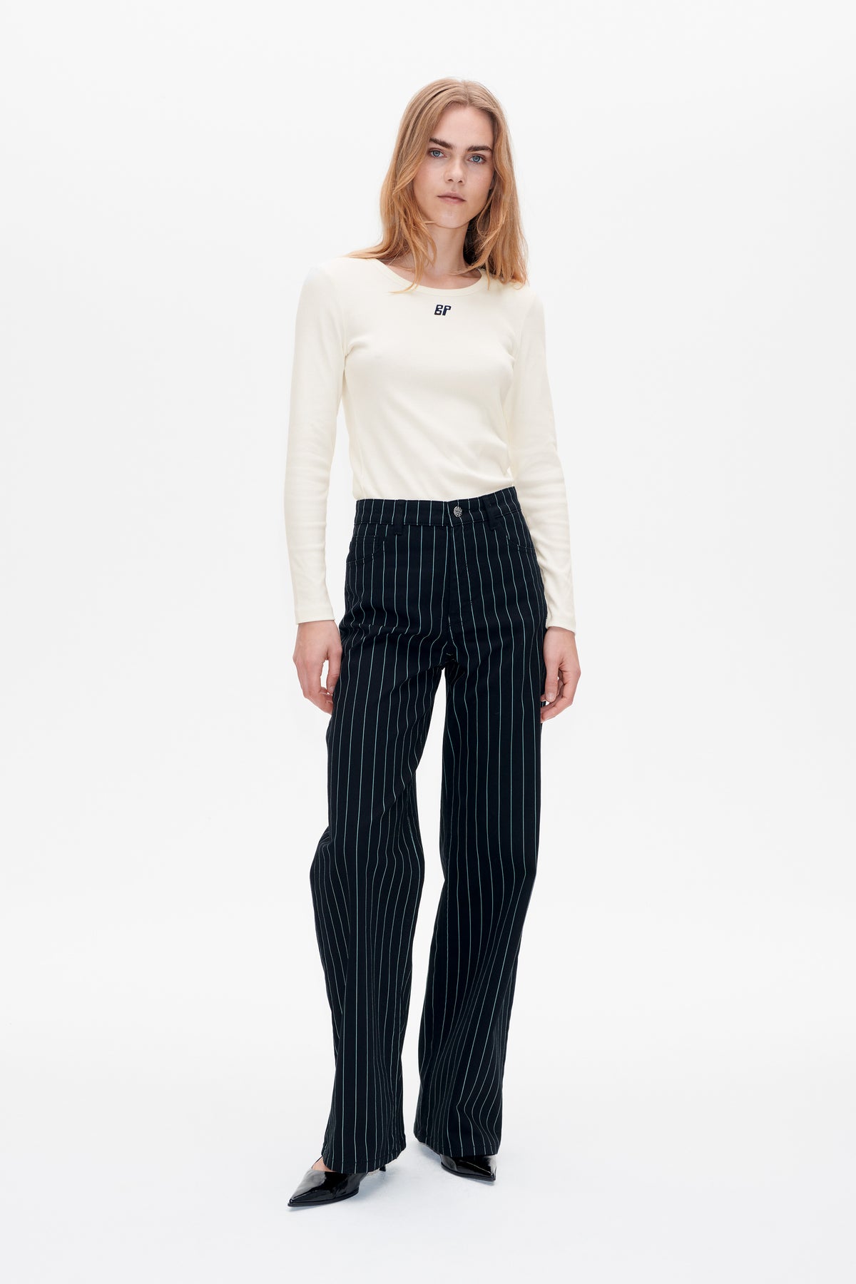 Frontale Studioaufnahme eines Looks mit elfenbeinfarbenem Langarmtop mit kleinem Brustlogo und einer marineblauen, weit geschnittenen Hose mit feinen Längsstreifen.