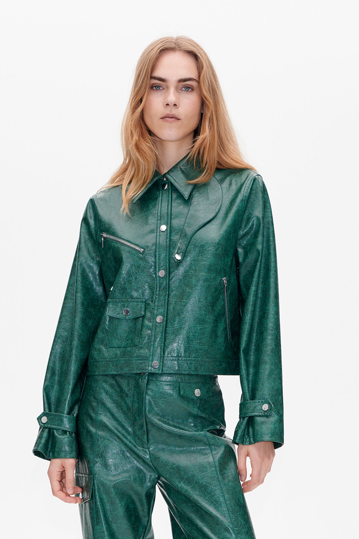 Front view of a model wearing a glossy, distressed dark green jacket with a pointed collar, snap buttons, zip pockets and flap pockets.