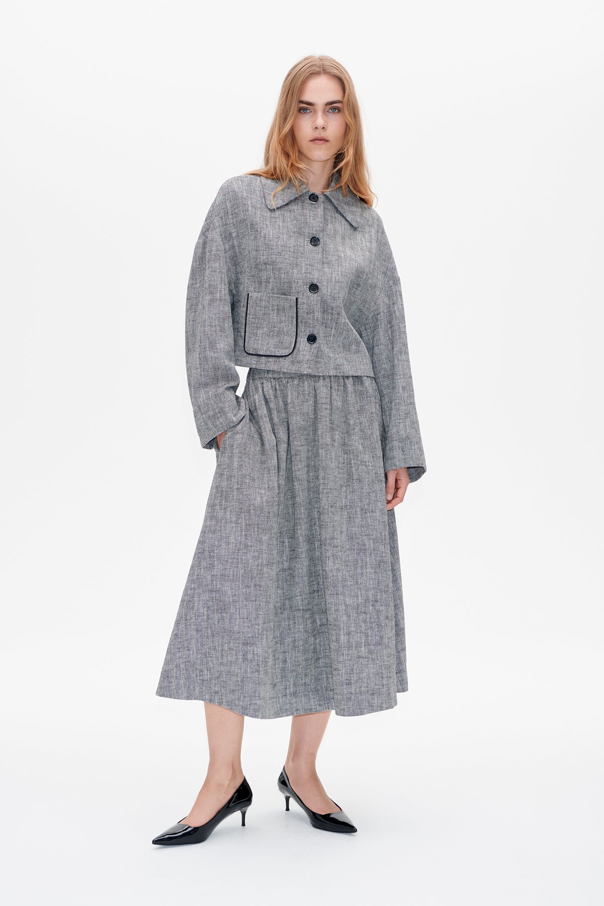 Model trägt eine graue Jacke mit Fischgrätmuster, Knopfleiste und großer paspelierter Tasche sowie einen passenden plissierten Midi-Rock.