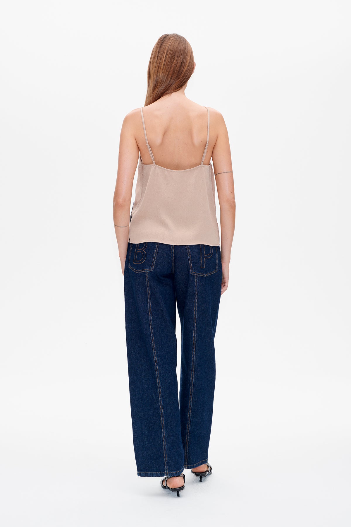 Rückenansicht eines Models in einem beigefarbenen Top mit dünnen Trägern und dunkelindigofarbenen, weit geschnittenen Jeans mit groß umrissenen Schriftzügen auf den Gesäßtaschen.
