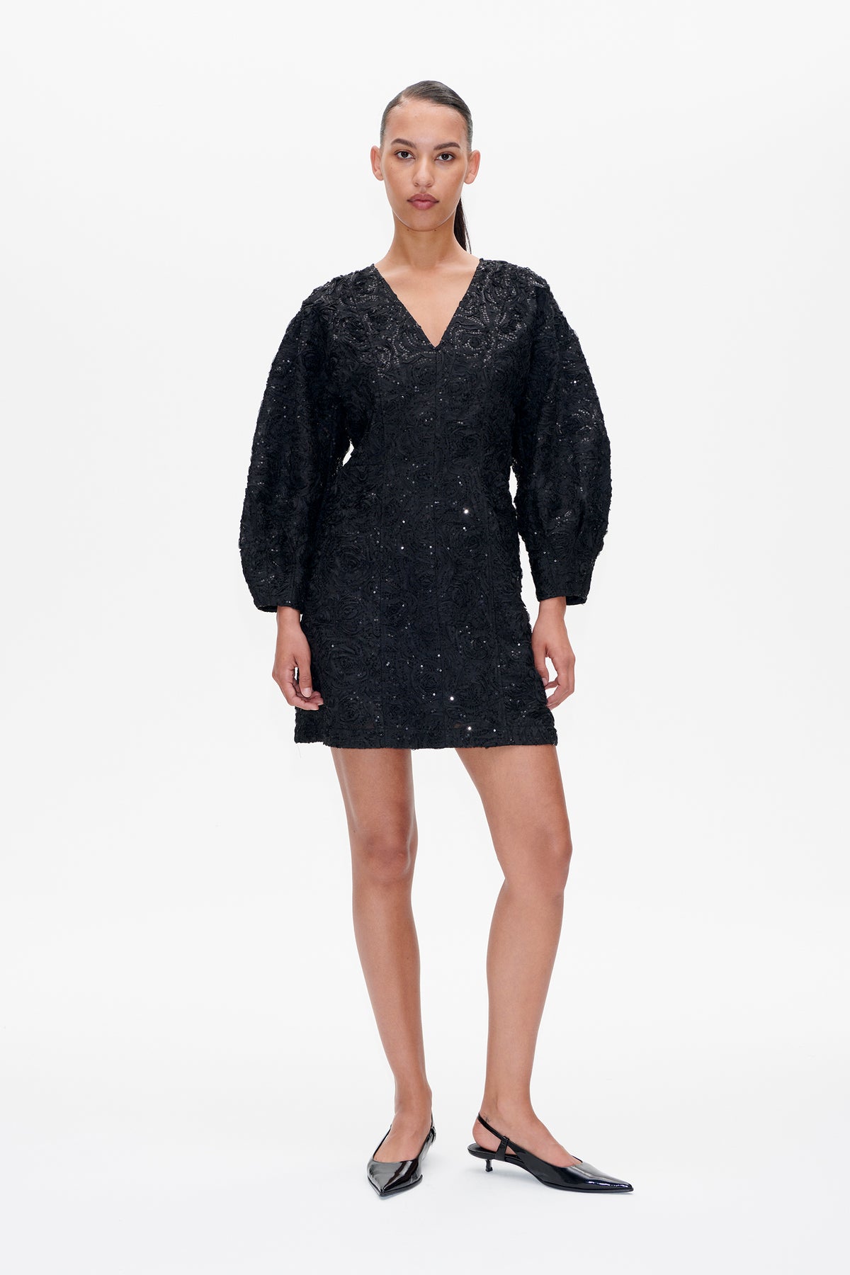 Model wearing a black V-neck mini dress with voluminous long sleeves, textured floral embroidery and scattered sequins, shown from the front.
