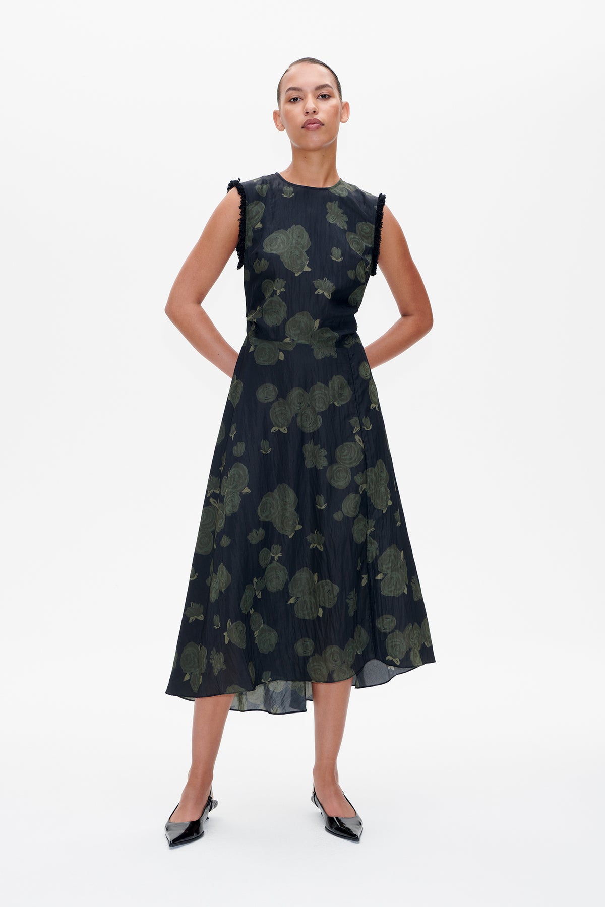Front view of a model wearing a dark sleeveless midi dress with a muted green floral print and frayed shoulder edges.