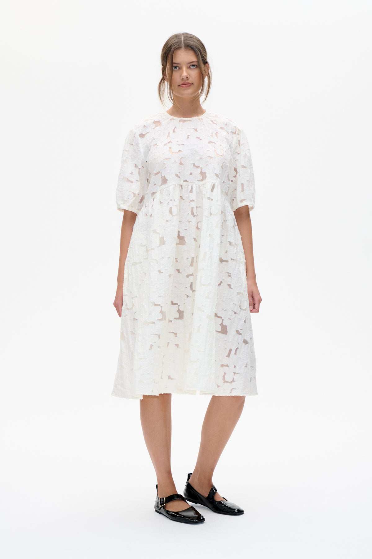 Model wearing a white knee-length dress with a round neckline, elbow-length sleeves and textured floral lace, shown from the front.