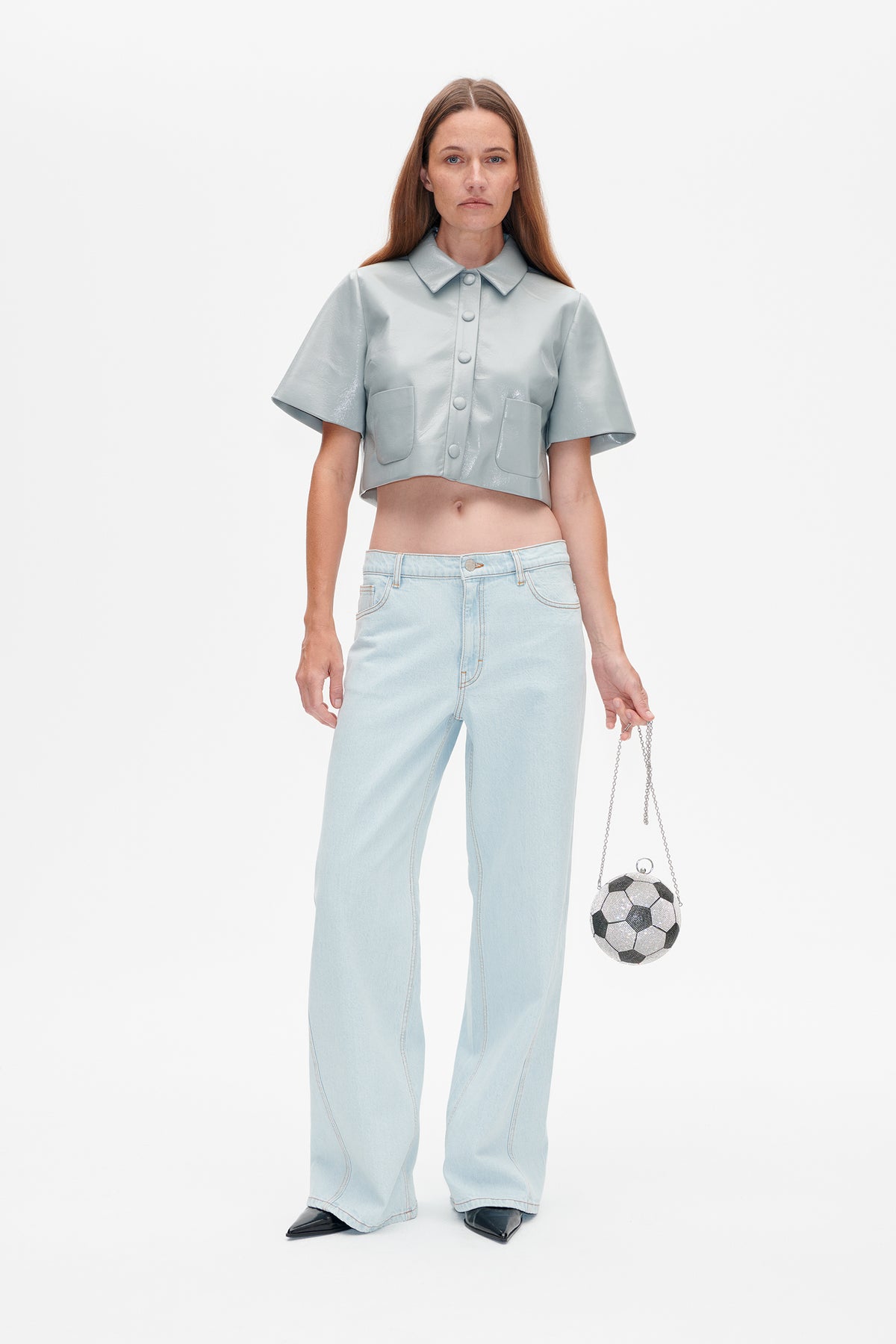 Full-length studio view of a model in a pale blue glossy cropped shirt with patch pockets, light-wash wide-leg jeans and a soccer-ball-shaped bag.