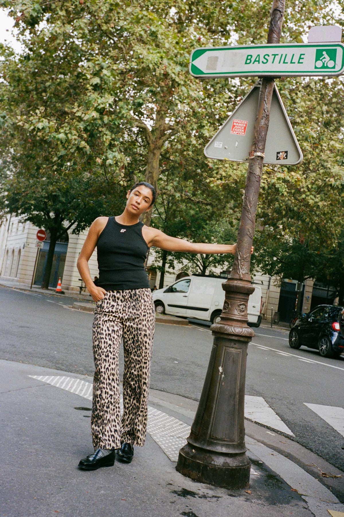 A model poses beside a Bastille street sign in a dark sleeveless top and loose beige trousers with a black leopard print.