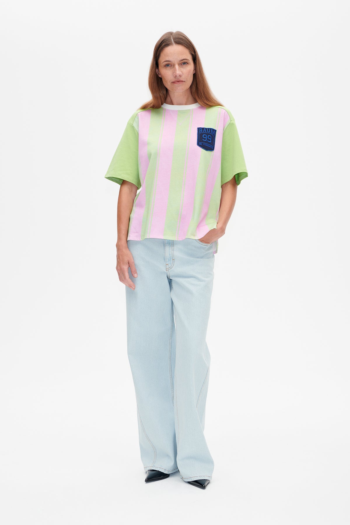 Model trägt ein locker sitzendes, kurzärmeliges T-Shirt mit vertikalen rosa- und hellgrünen Streifen, unifarbenen grünen Ärmeln und einem blauen Brustaufnäher.