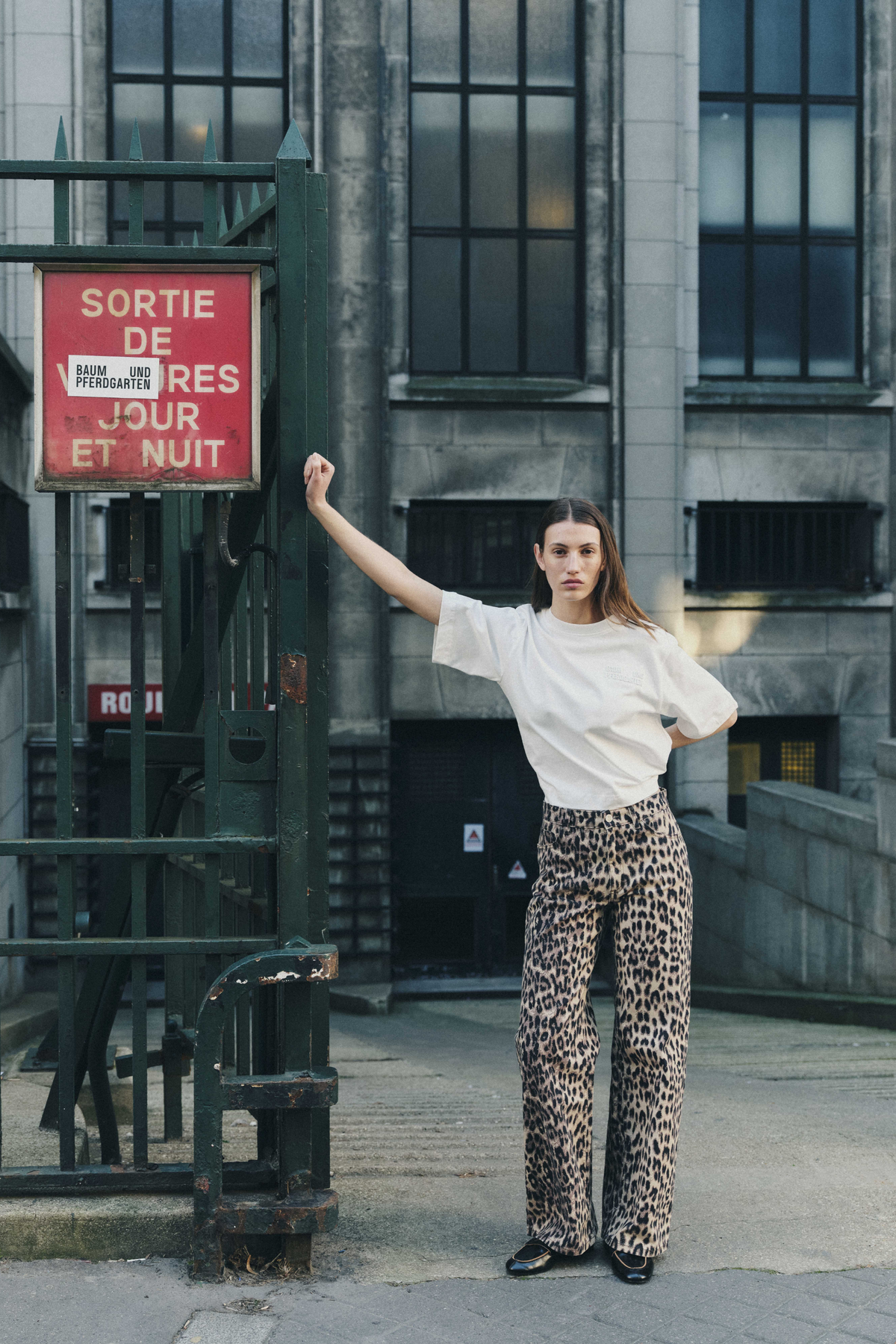 Model poses beside a dark metal gate in a white oversized T-shirt and beige-and-black leopard-print trousers.