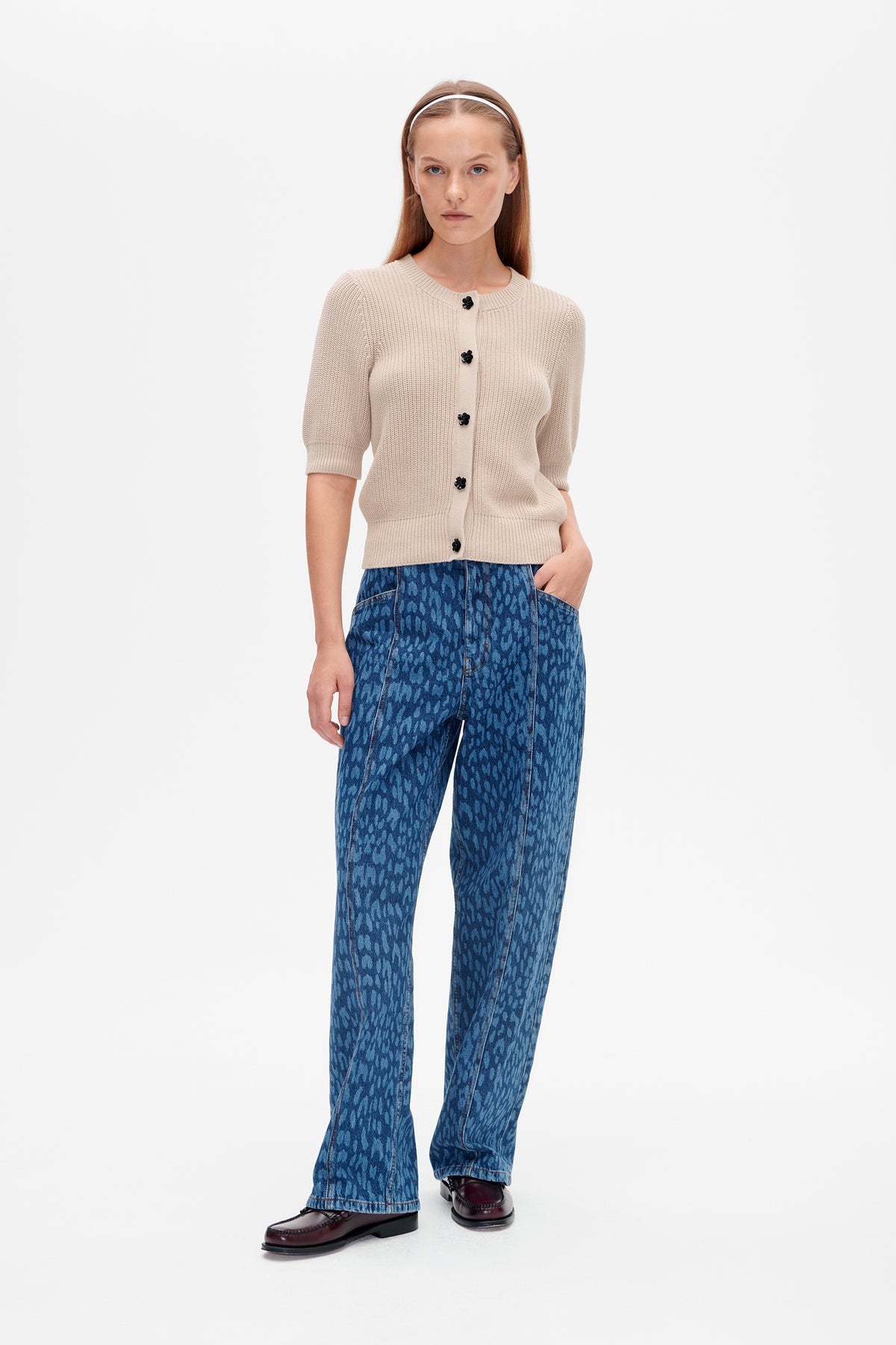 Model wearing a cream short-sleeved knit cardigan with black flower-shaped buttons and blue patterned jeans, shown from the front.
