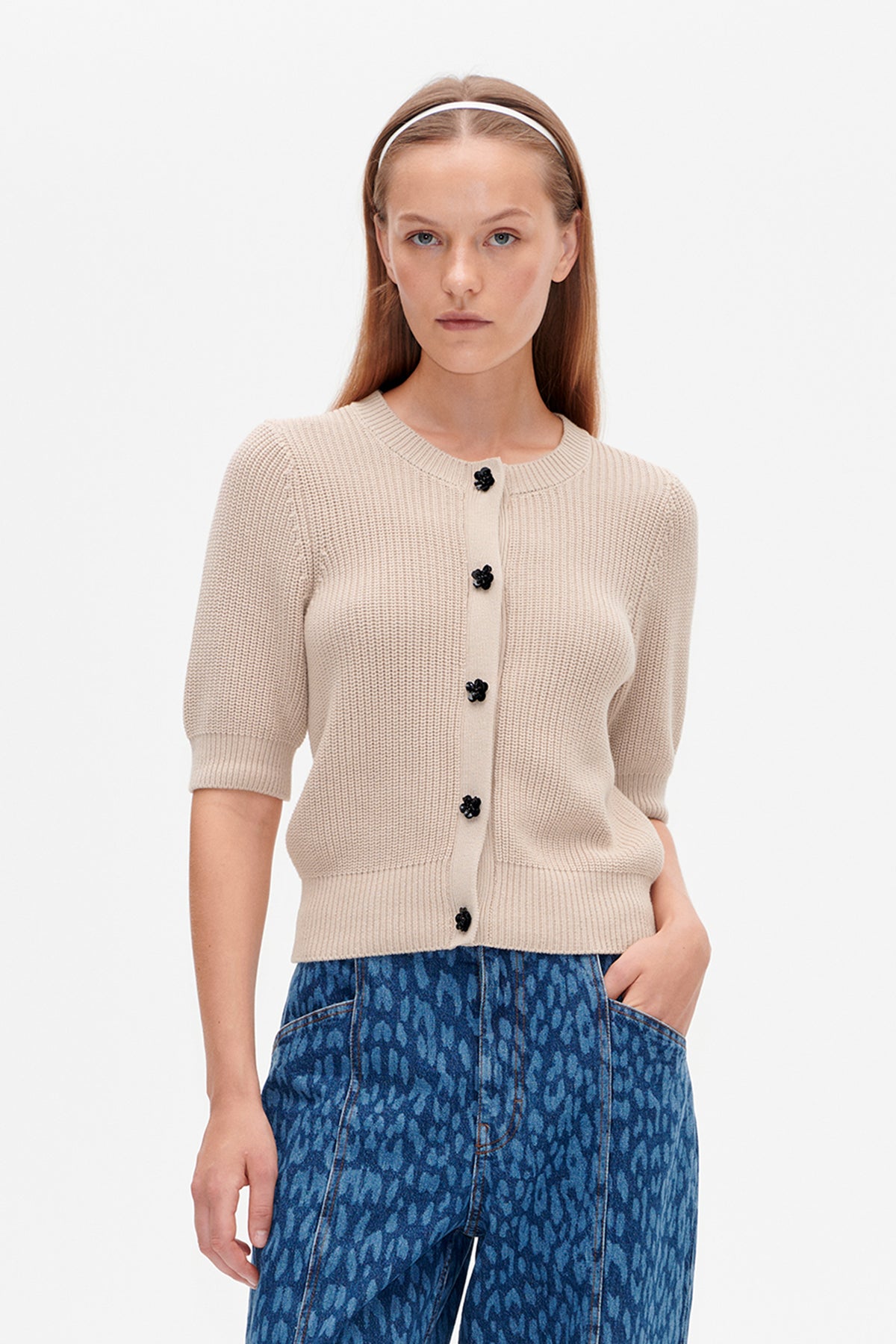 Front view of a model wearing a cream ribbed knit cardigan with black floral buttons and blue patterned jeans.