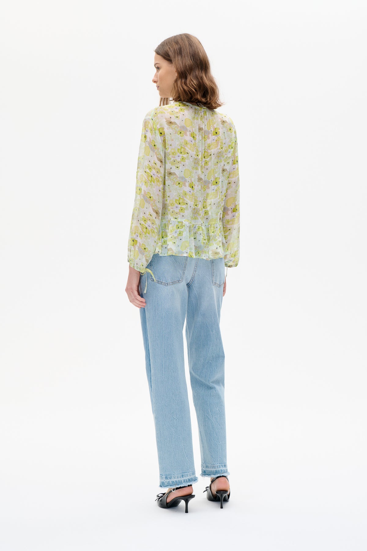Model viewed from behind wearing a sheer pale blouse with a yellow-green floral print, long sleeves and a gathered peplum hem, with light blue jeans.