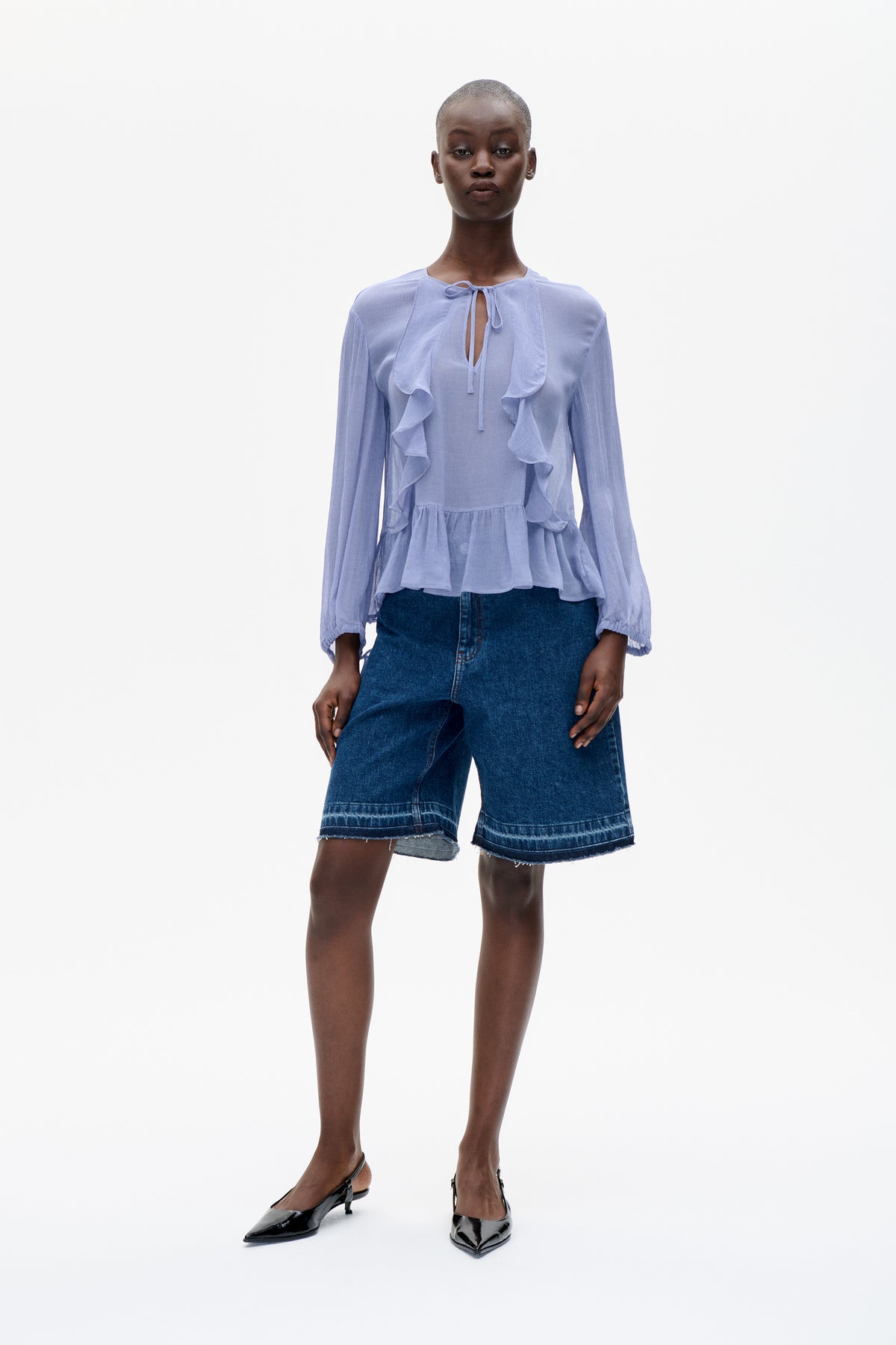 Model wearing a pale lavender long-sleeved blouse with a tie neckline and cascading ruffles, paired with dark denim shorts.