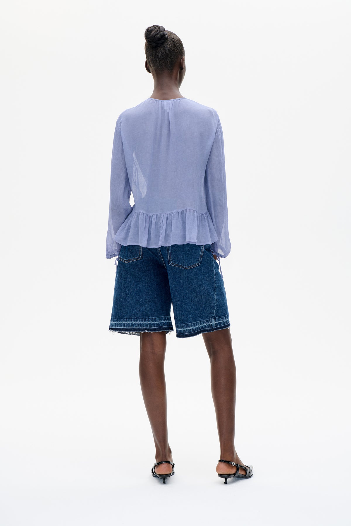 Back view of a pale lavender long-sleeved blouse with a gathered peplum hem, paired with dark denim shorts.