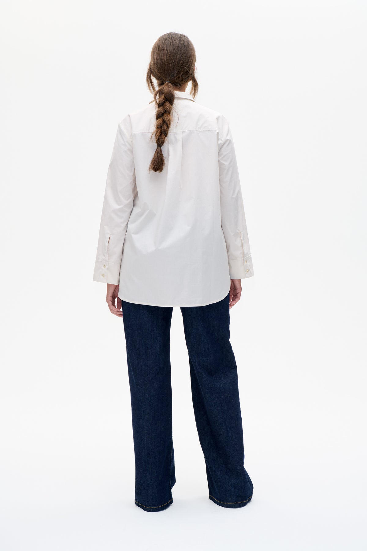 Back view of a white long-sleeved collared shirt with a rear yoke seam and curved hem, paired with dark wide-leg jeans.