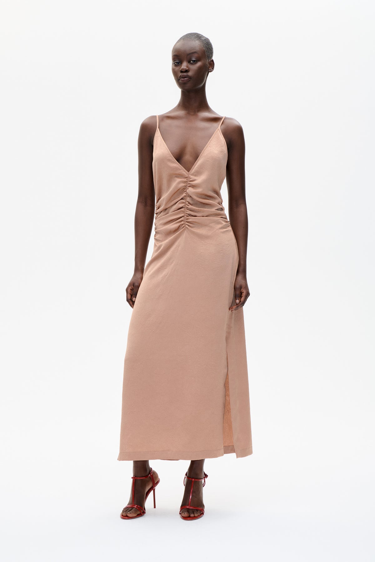 Front view of a model wearing a peach-pink midi dress with thin straps, a deep V-neck, and gathered ruching down the front.