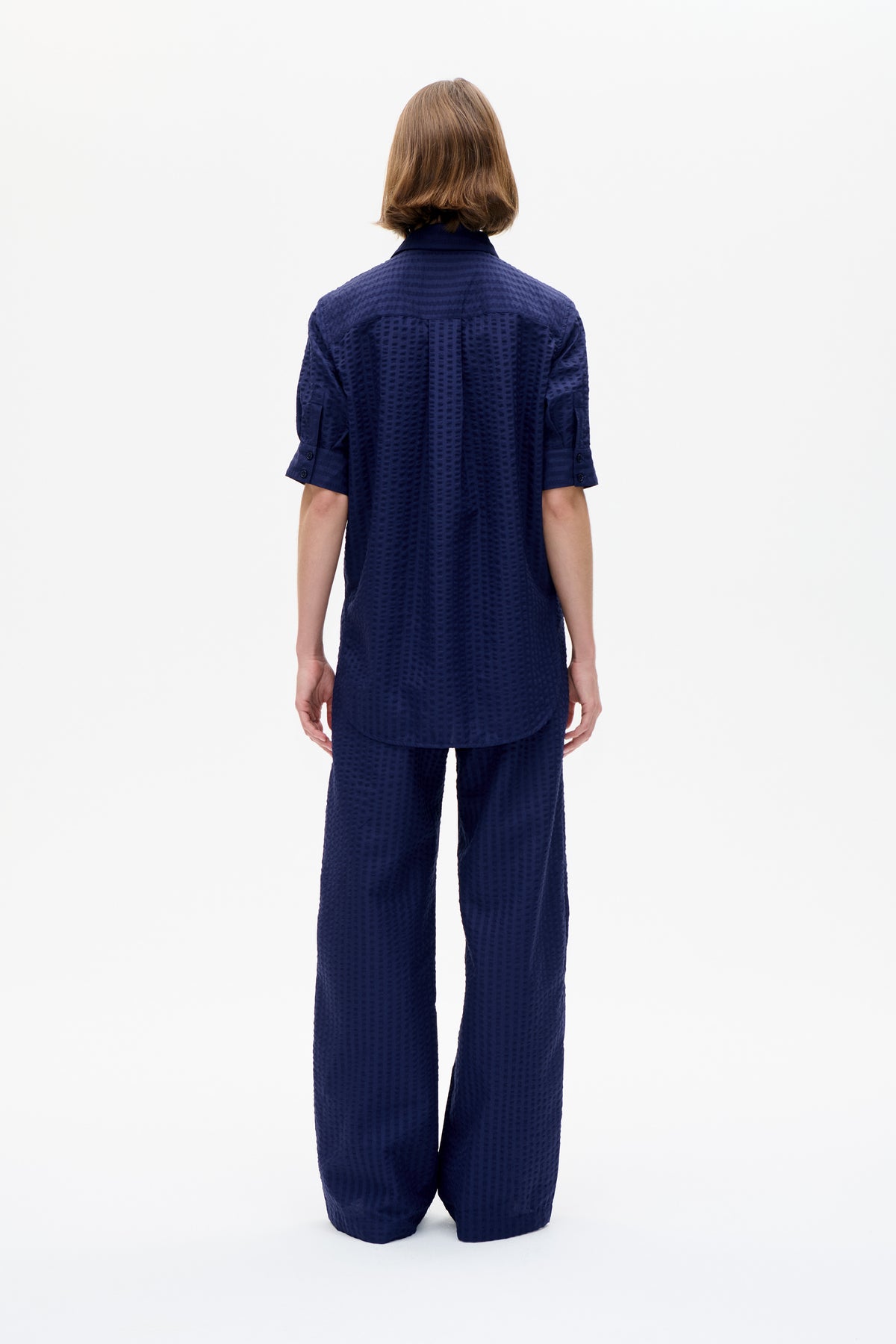 Vue de dos d'un mannequin portant une chemise bleu marine à col et manches courtes, et un pantalon à coupe large assorti à motif discret en relief.