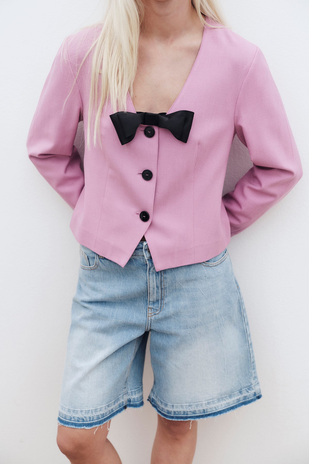 Campaign close-up of a model wearing a pink tailored blazer with a black bow and buttons, paired with light-wash denim shorts.
