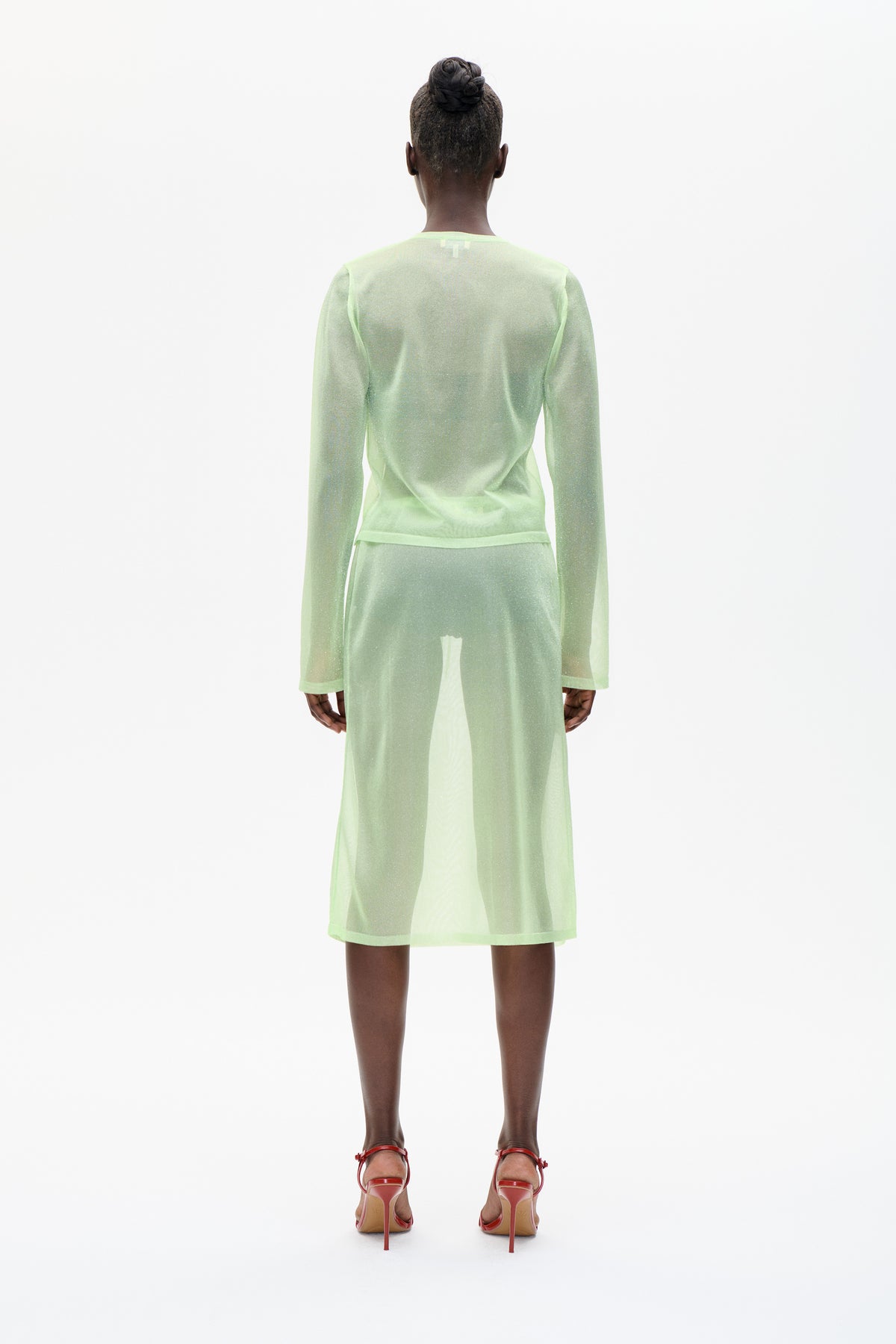 Rear view of a model wearing a sheer, subtly shimmering pale lime-green long-sleeved top and matching midi skirt.