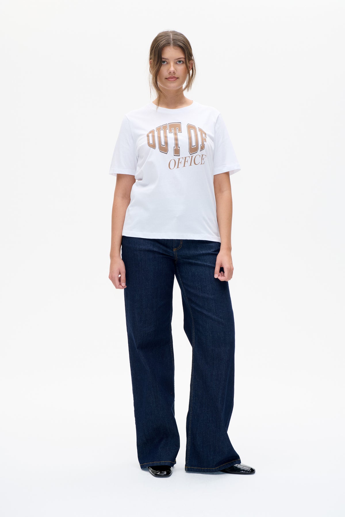 Vue de face d'un mannequin portant un T-shirt blanc à manches courtes avec l'inscription beige "OUT OF OFFICE" et un jean large foncé.