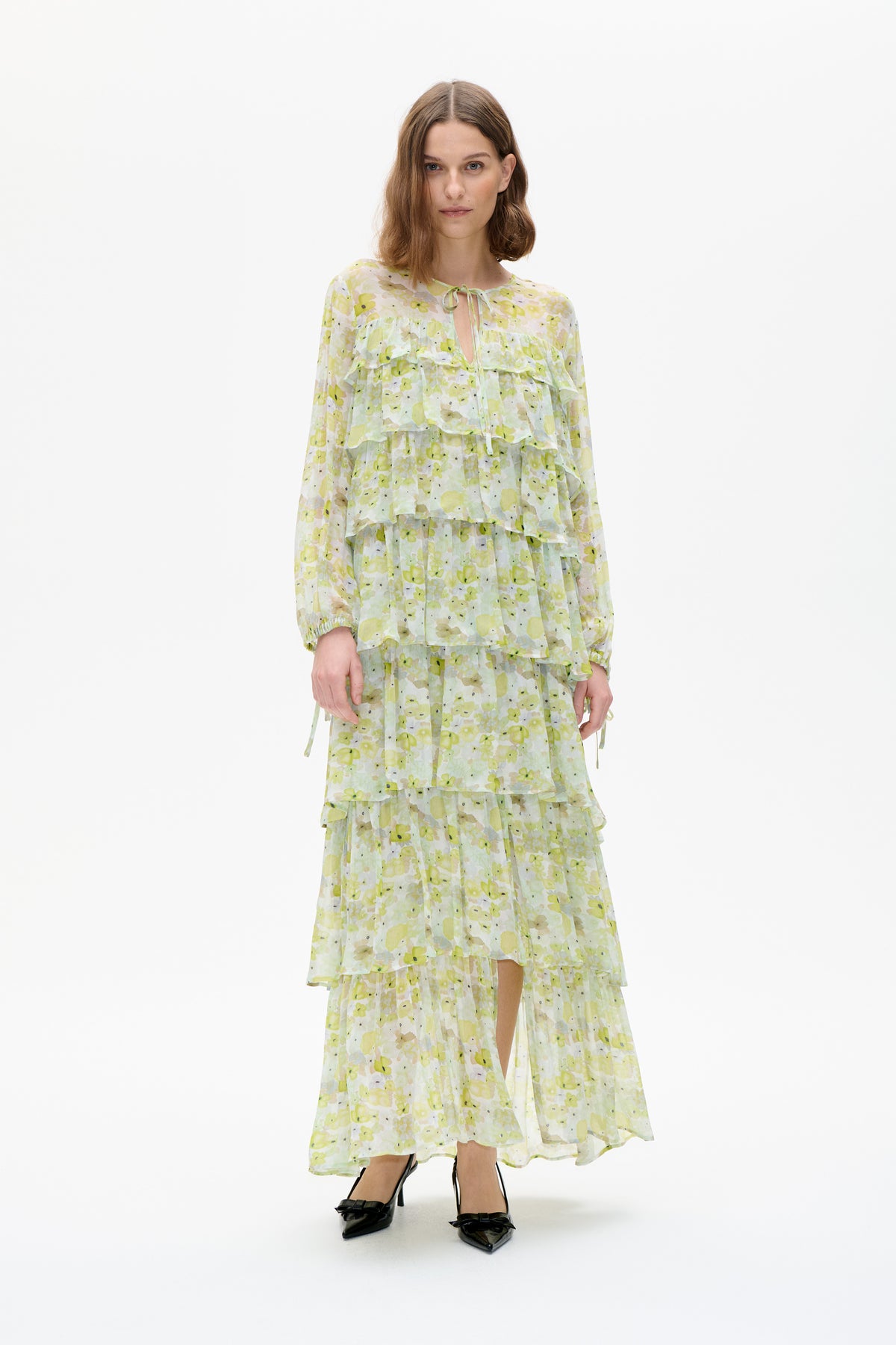 Model wearing a layered, long-sleeved dress with a pale yellow-green floral print, ruffled tiers and a tie neckline.