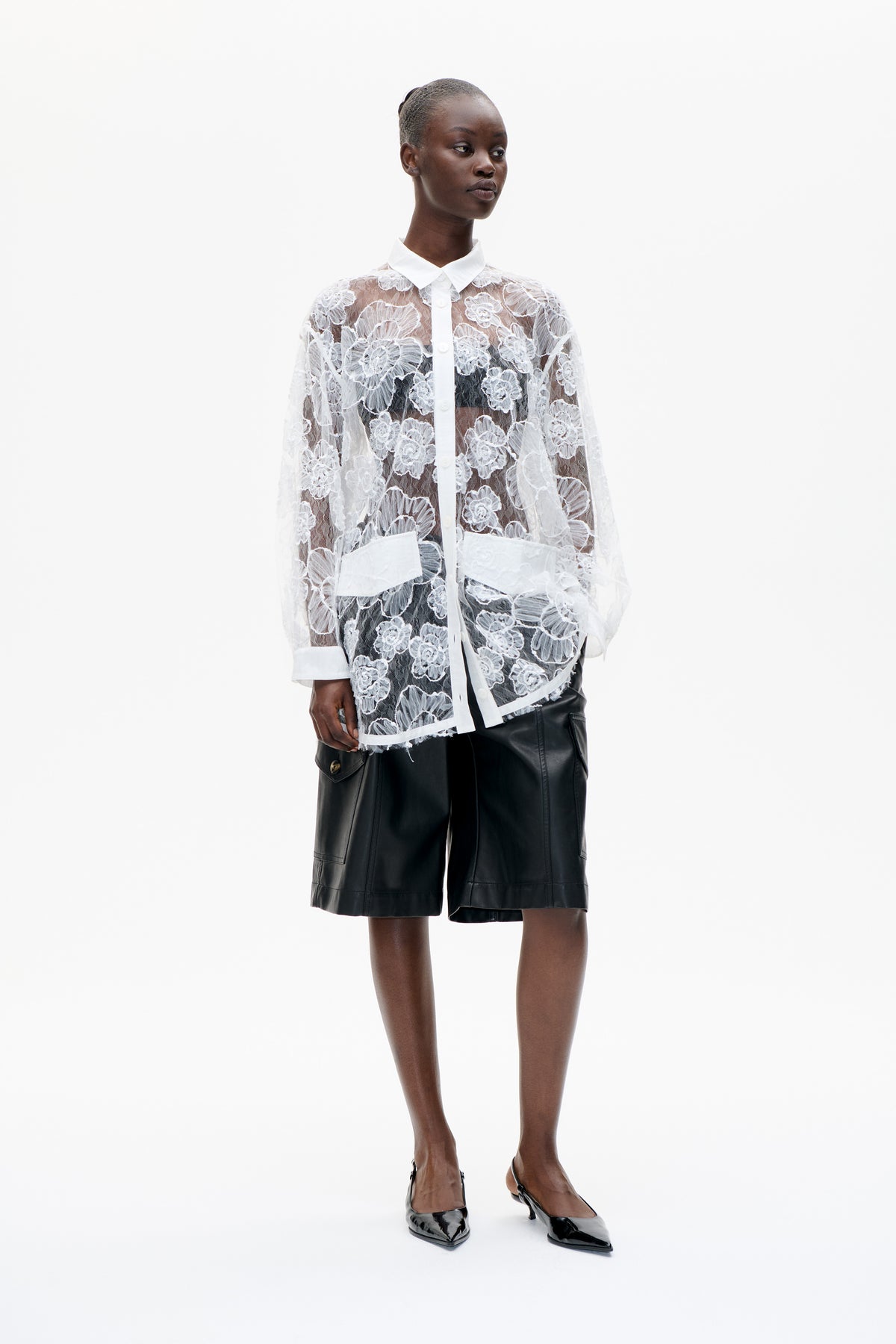 Model wearing a sheer white floral lace button-front shirt with a pointed collar and black knee-length shorts against a white background