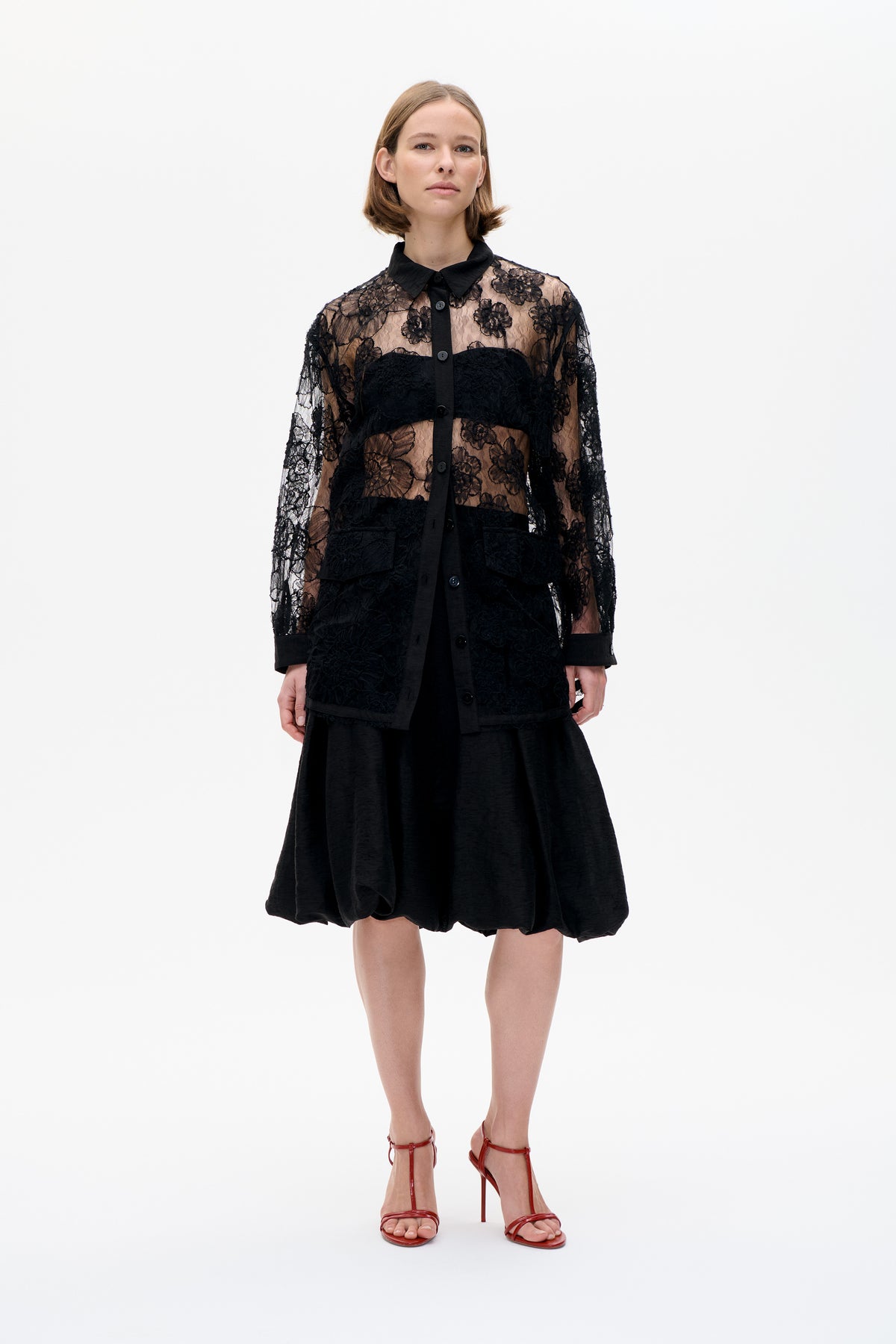 Front view of a model wearing a sheer black floral lace button-front shirt with solid panels over a black balloon-hem dress.