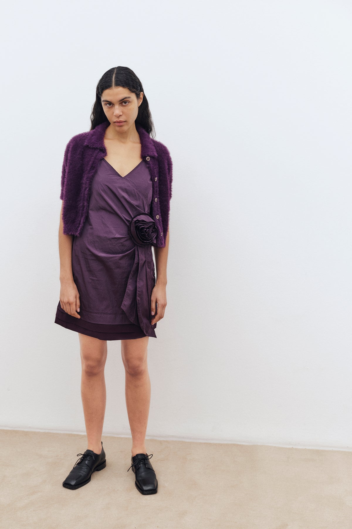 A model wears a plum fuzzy button-front cardigan over a draped purple V-neck mini dress with a large fabric flower at the waist.