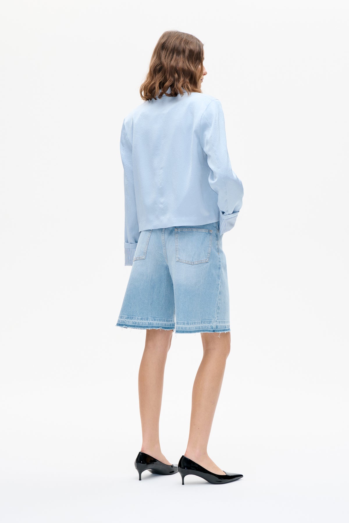Back view of a model wearing a pale blue textured long-sleeve shirt with striped cuffs and light-wash denim shorts.