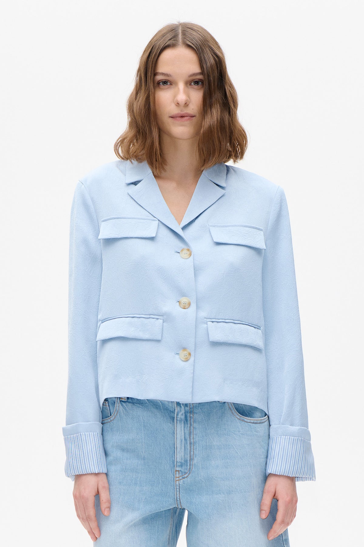 Front view of a light blue textured blazer with large flap pockets, pale buttons and striped turned-up cuffs, worn with light-wash jeans.