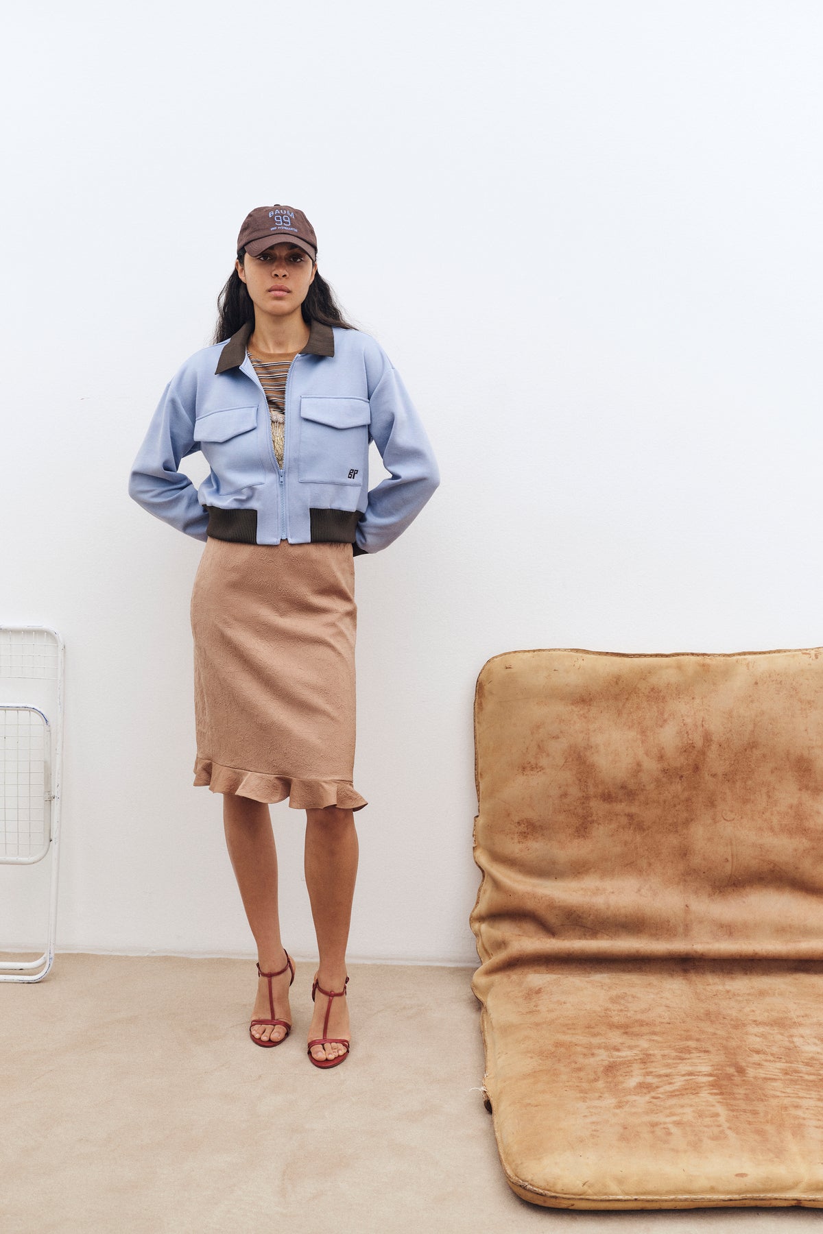 Model tegen een lichte muur draagt een lichtblauwe jas met ritssluiting en bruine bies, een gestreepte top, een taupekleurige midirok met structuur en een bruine pet.