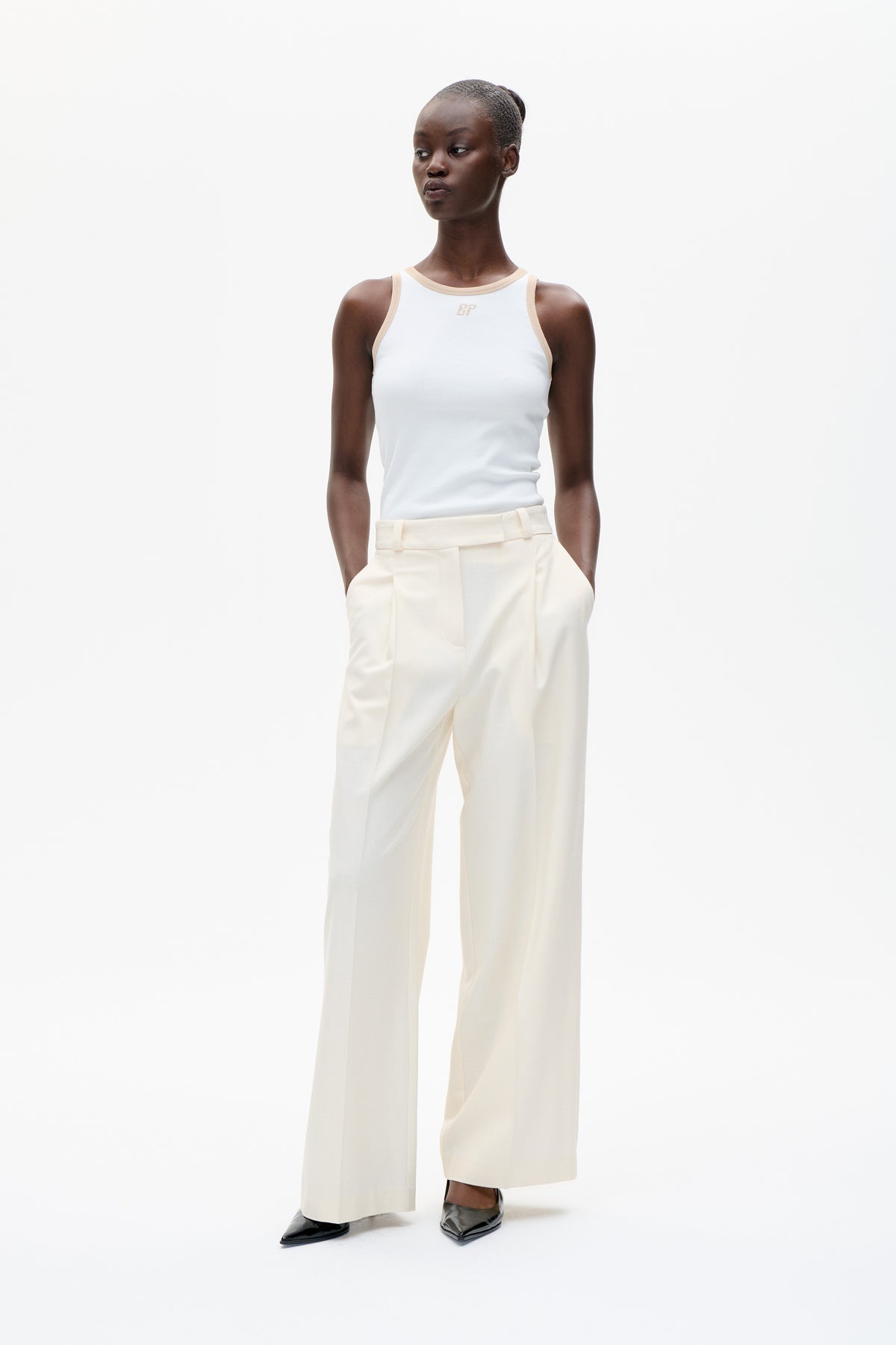 Model trägt ein weißes, ärmelloses Top mit beiger Einfassung und kleinem Brustmonogramm, dazu eine cremefarbene, weite Hose.