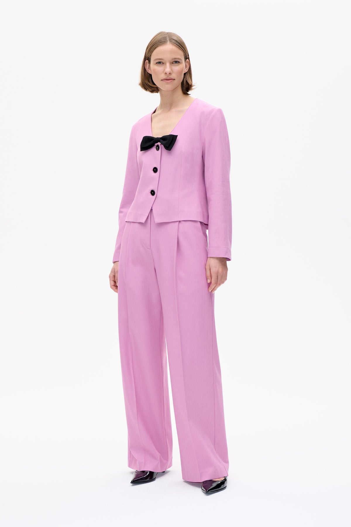 Model trägt eine pinke Jacke mit Knopfleiste und schwarzem Schleifendetail sowie passende, weit geschnittene Hosen. Aufnahme von vorne.
