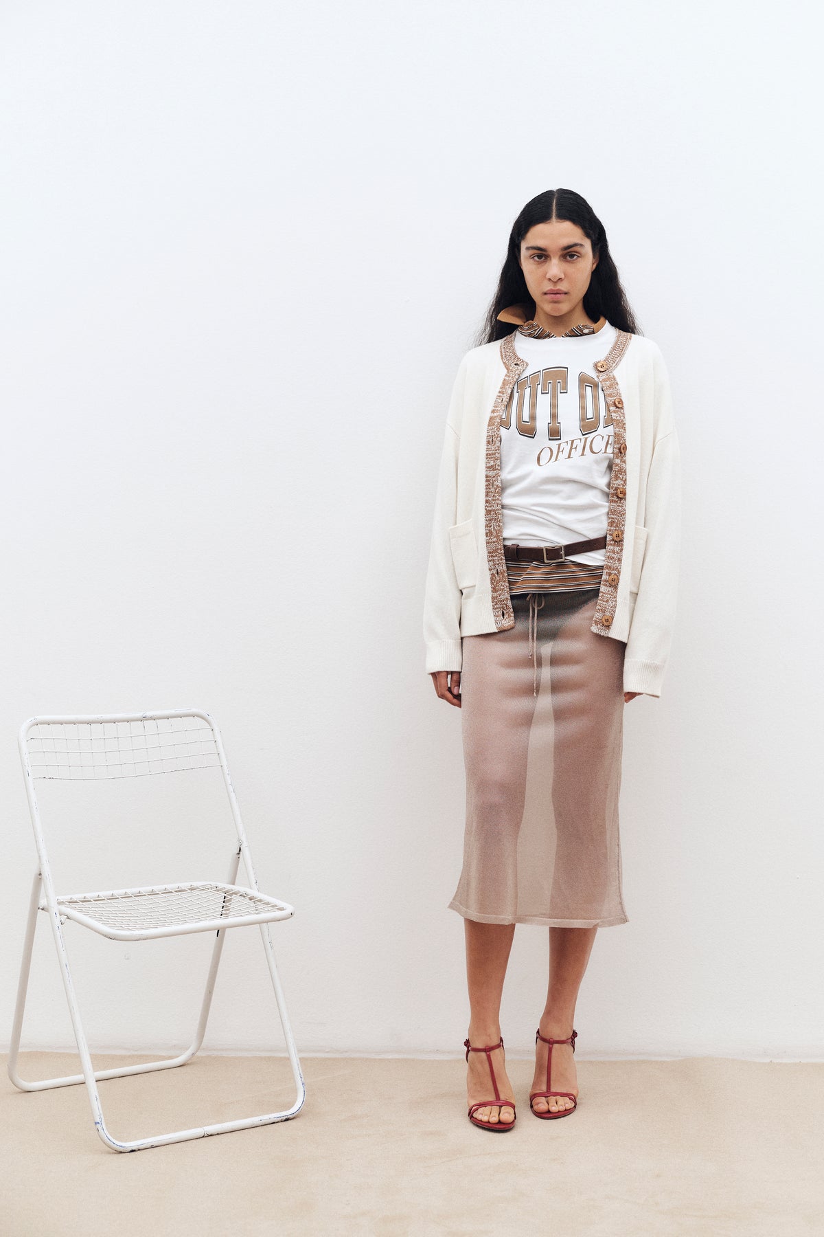 Campaign portrait beside a white chair, featuring an ivory cardigan with brown trim, an “OUT OF OFFICE” graphic tee and a sheer beige midi skirt.