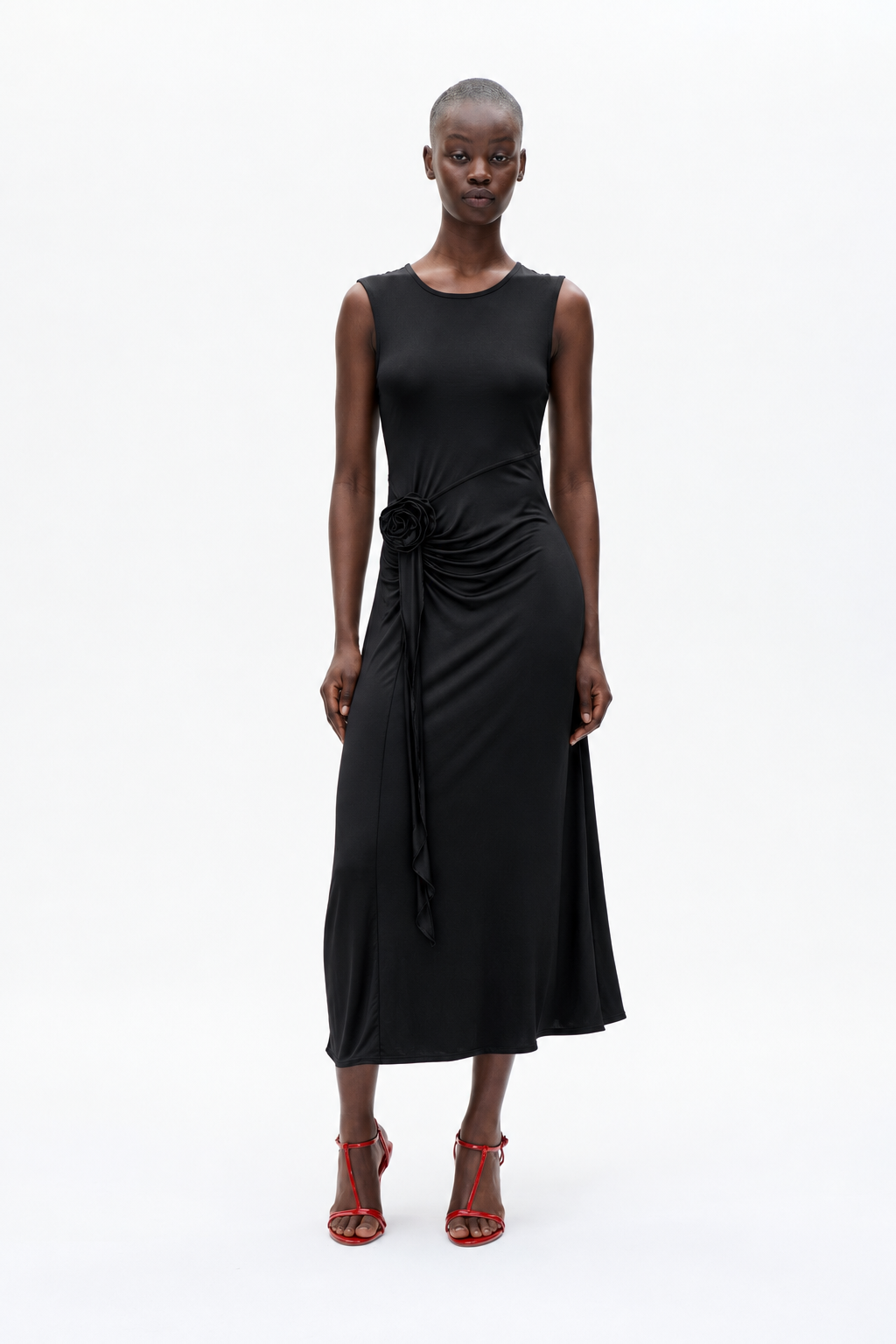 Model wearing a sleeveless black midi dress with a round neckline, gathered waist, and fabric rose detail at the hip.